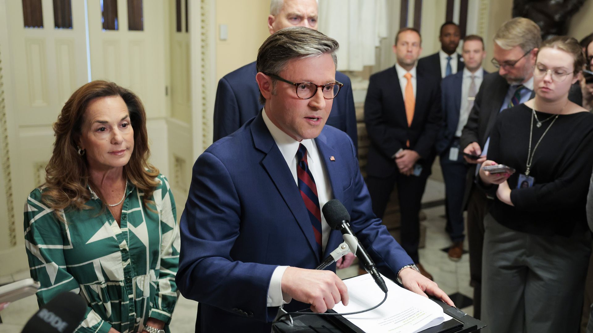 Man in a blue suit speaks at a podium with a microphone and papers, flanked by a woman in a green patterned dress; reporters observe in the background.