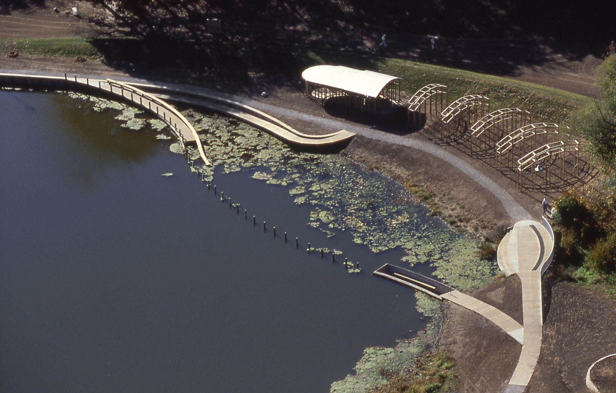 Aerial view of a park by a lake: curved wooden ramp, a sheltered pavilion over water, and multiple arched trellis structures along a curved gravel path, with green banks and lily pads.