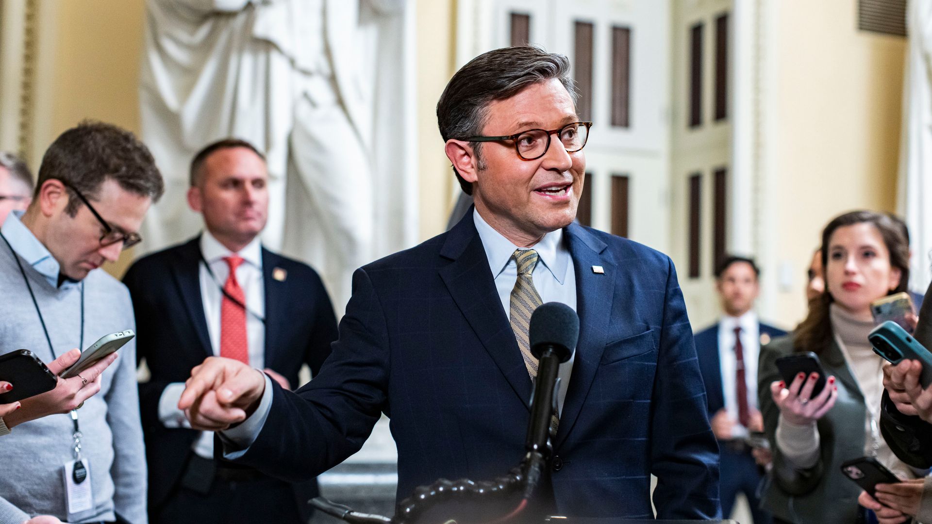 House Speaker Mike Johnson in a blue suit, speaking into a microphone surrounded by reporters.