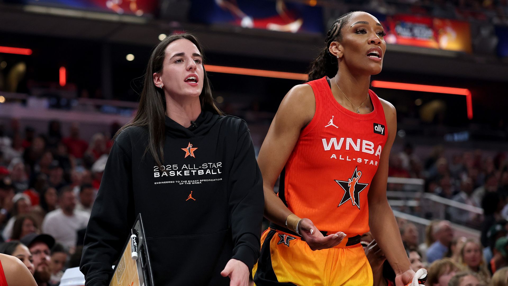 Two women walk the bench at a basketball event: on the left, a trainer in a black hoodie holding a clipboard; on the right, a player in an orange WNBA All-Star uniform, crowd in the stands.