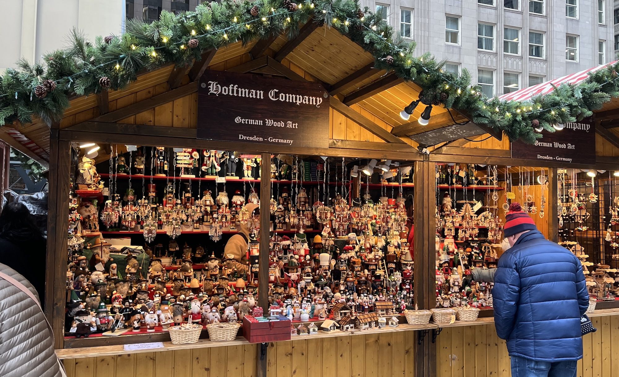 Wooden market stall decorated with garlands and lights, selling German wood art including nutcrackers and ornaments, with a man in a blue jacket and red hat browsing.