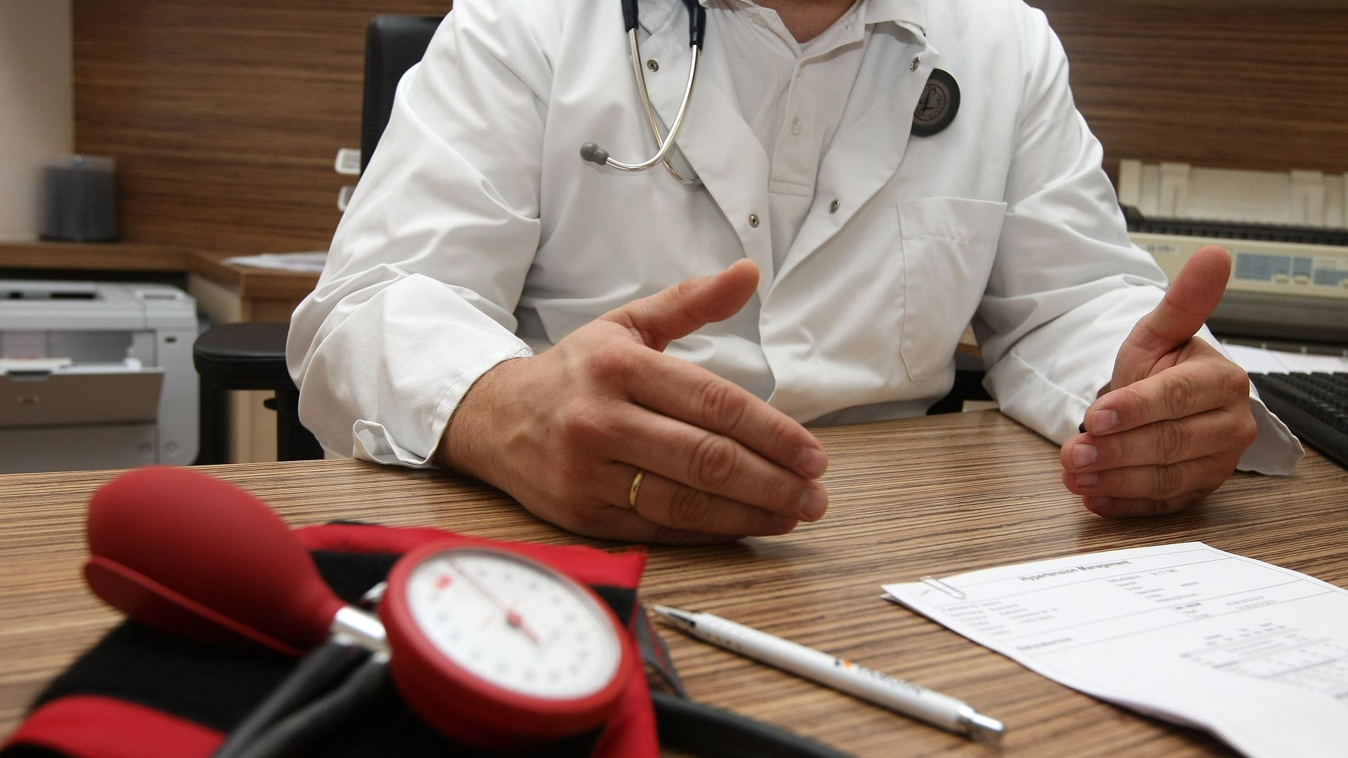 A doctor sits at a desk with paperwork and a blood pressure cuff.