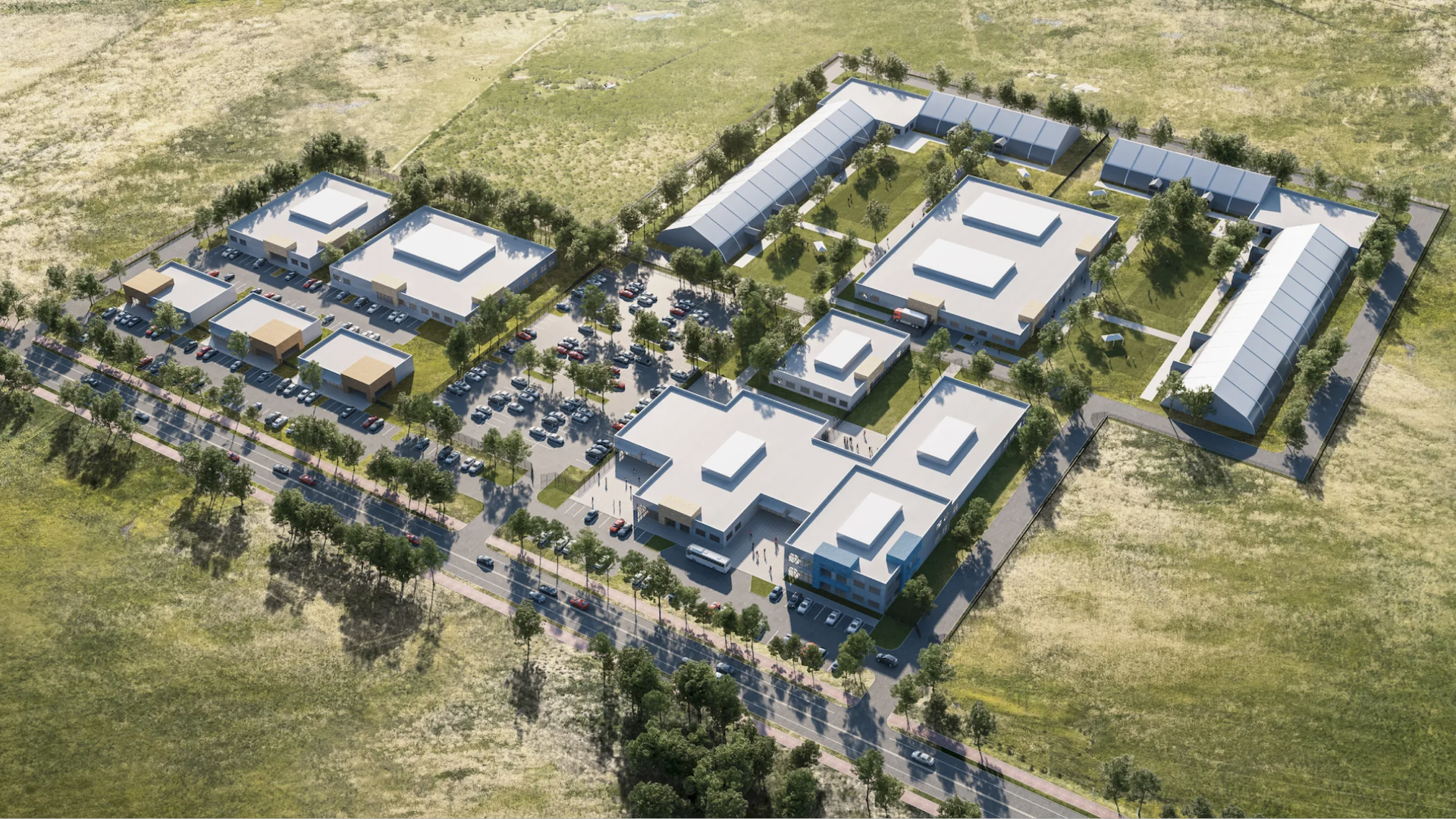Aerial view of a large campus with multiple white-roofed buildings, green lawns, trees, parking lots with cars, and surrounding grassy fields under daylight.