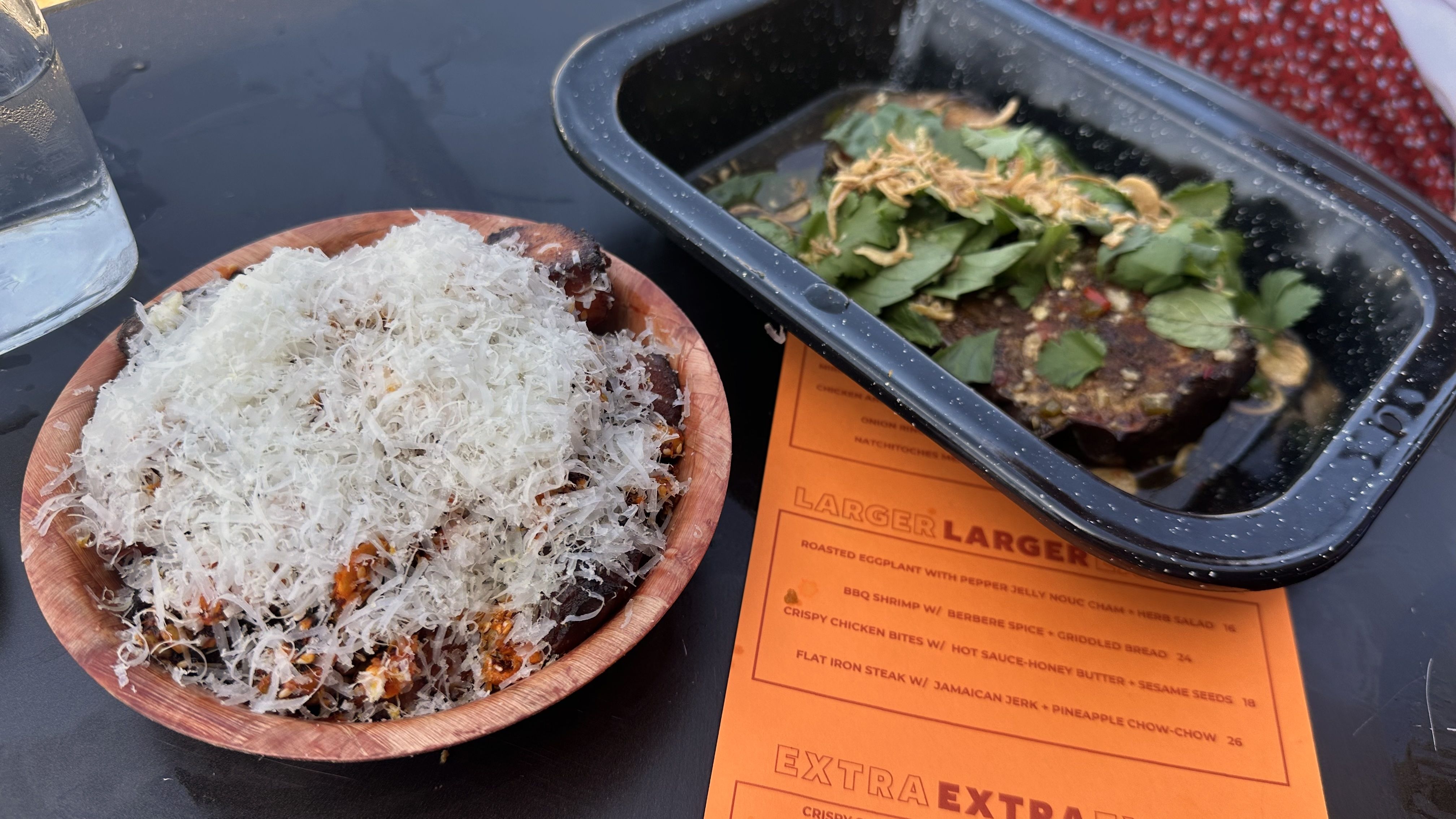 A round plate of food is covered with shaved pecorino, and at right, an enamel metal tin is filled with roasted eggplant covered with cilantro. Both sit on a black tabletop.