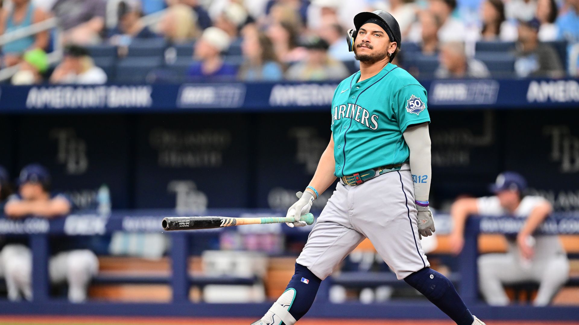 A baseball player in a teal jersey looks disappointed after batting. 