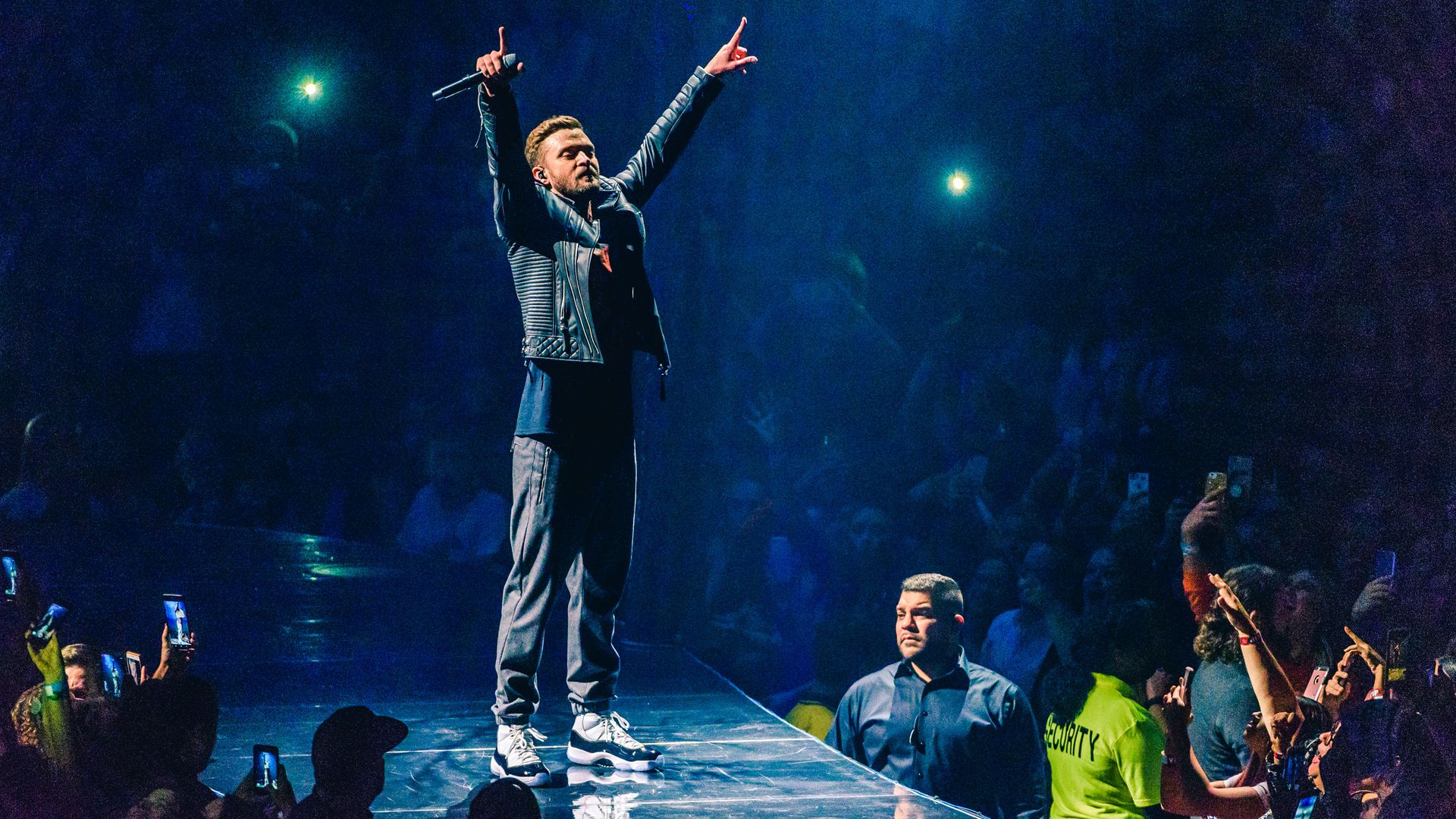 Justin Timberlake raises his hands on stage holding a microphone. 