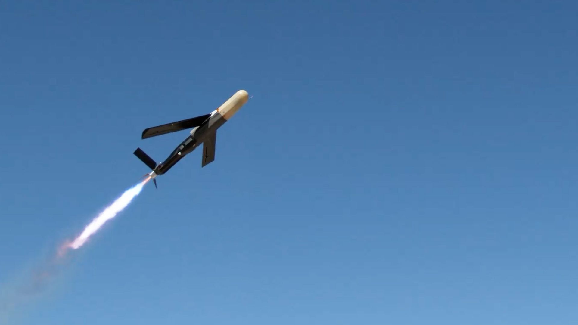 Drone with black wings and beige nose cone launching upward against a clear blue sky. It's emitting a bright white flame trail.