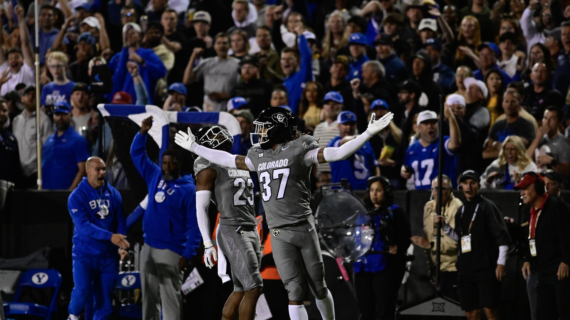 Two Colorado football players in gray uniforms, numbers 23 and 37, celebrating on the field with number 37 spreading arms wide while a crowd in blue cheers in the background.