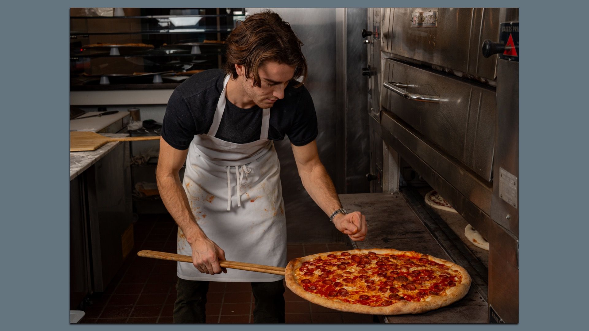 Mason Lesser in a black T-shirt and white apron leans over a wooden pizza peel, pulling a large pepperoni pizza from a stainless steel oven in a busy pizzeria kitchen.