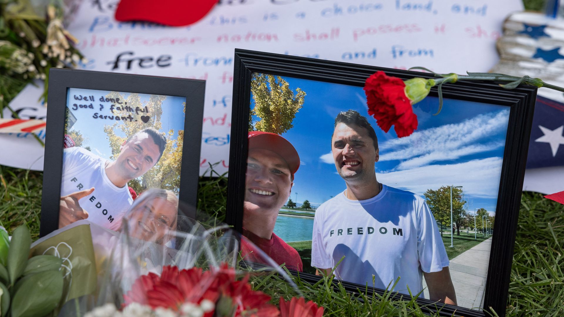 Memorial with framed photos of two men, one wearing a white shirt with FREEDOM, red carnations, flowers, an American flag, and a red "Make America Great Again" cap on the grass.
