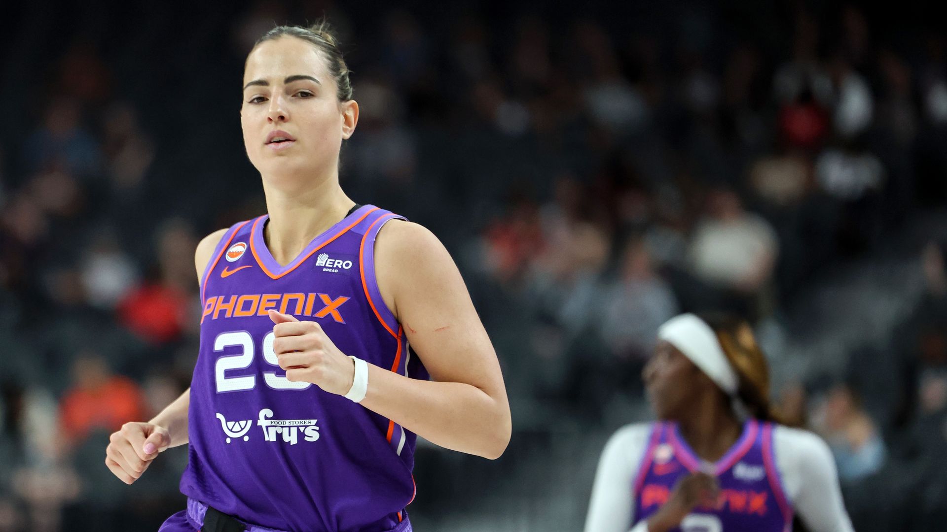 Female basketball player in a purple uniform with orange trim, number 23 and PHOENIX on the chest, running on the court with a blurred crowd in the background.