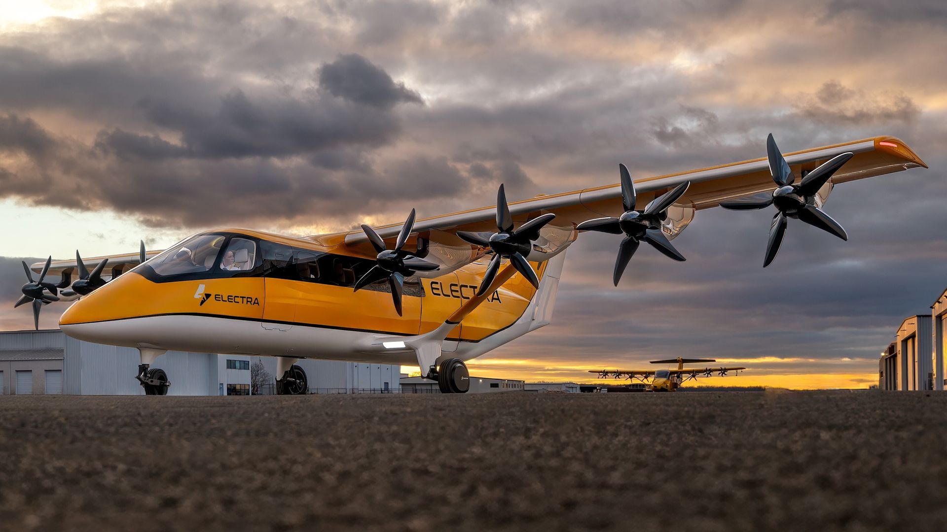Bright yellow Electra seaplane with multiple propellers along its wing sits on a tarmac beneath a dramatic cloudy sunset; hangar buildings line the background.