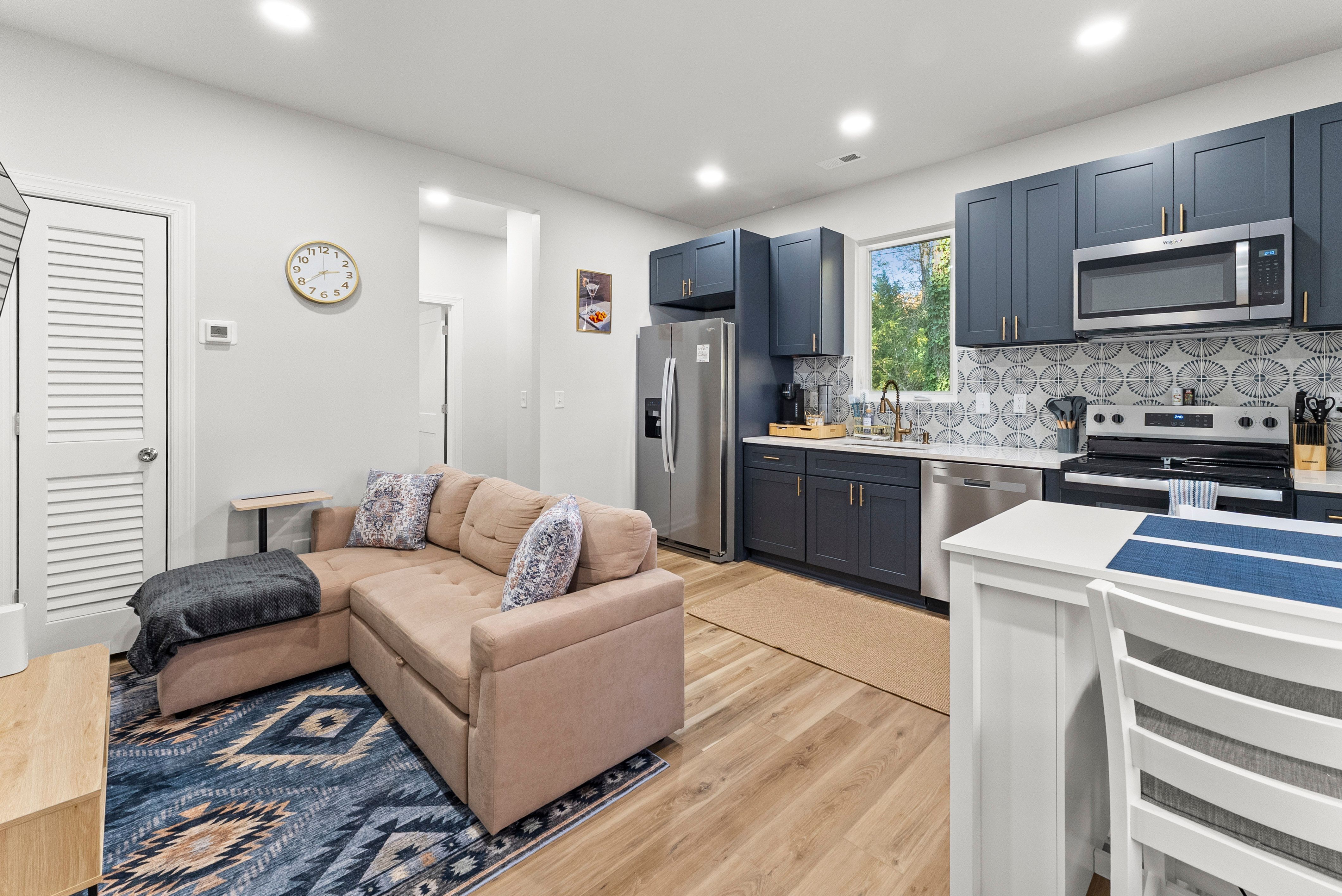 Open living area with beige L-shaped sofa on blue patterned rug, adjacent to modern kitchen with navy blue cabinets, stainless steel appliances, patterned backsplash, and window overlooking trees.