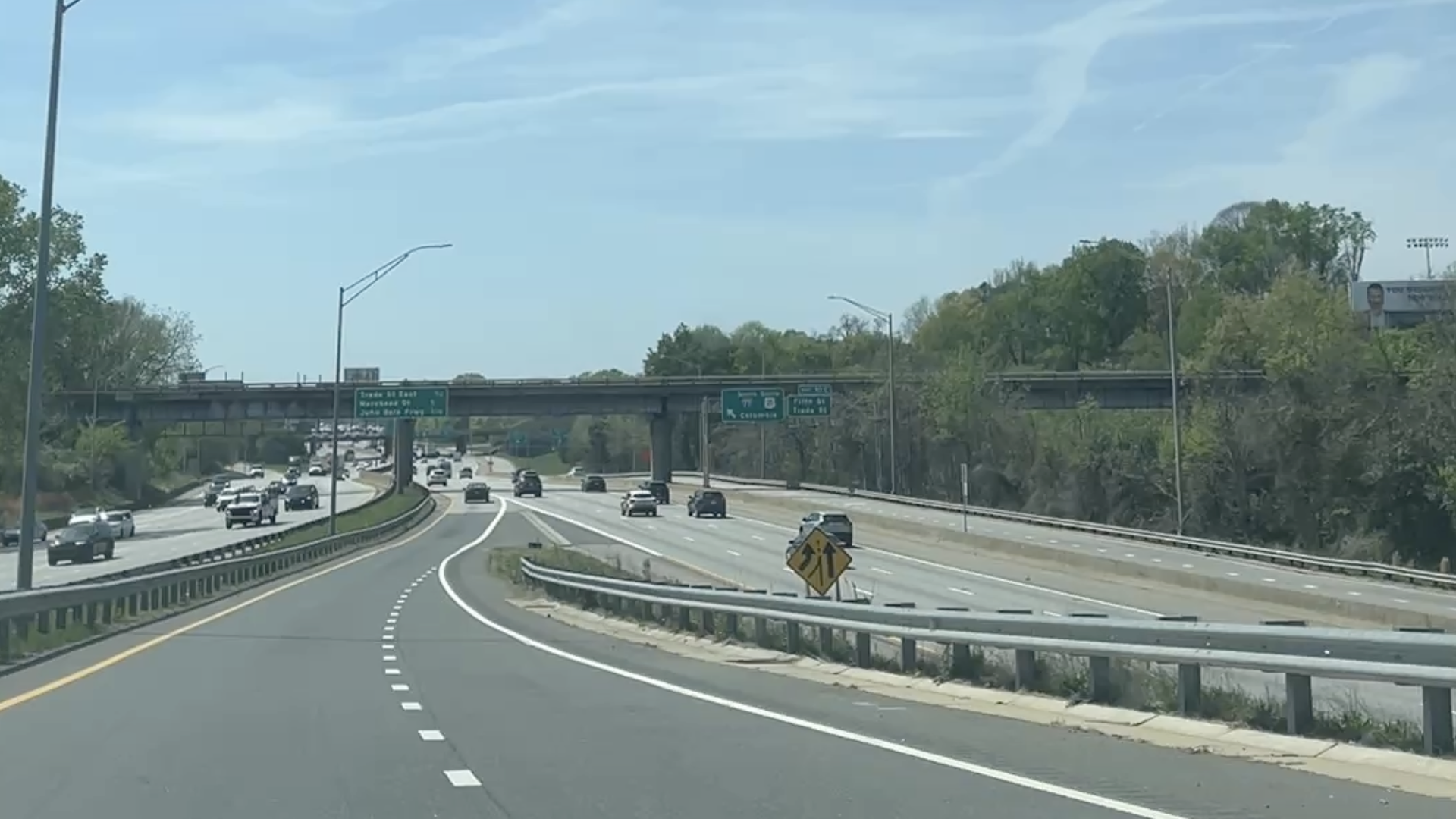 Multi-lane highway with an overpass and green exit signs. A yellow warning sign sits in the median; traffic travels in both directions while trees line the road shoulders under a clear blue sky.