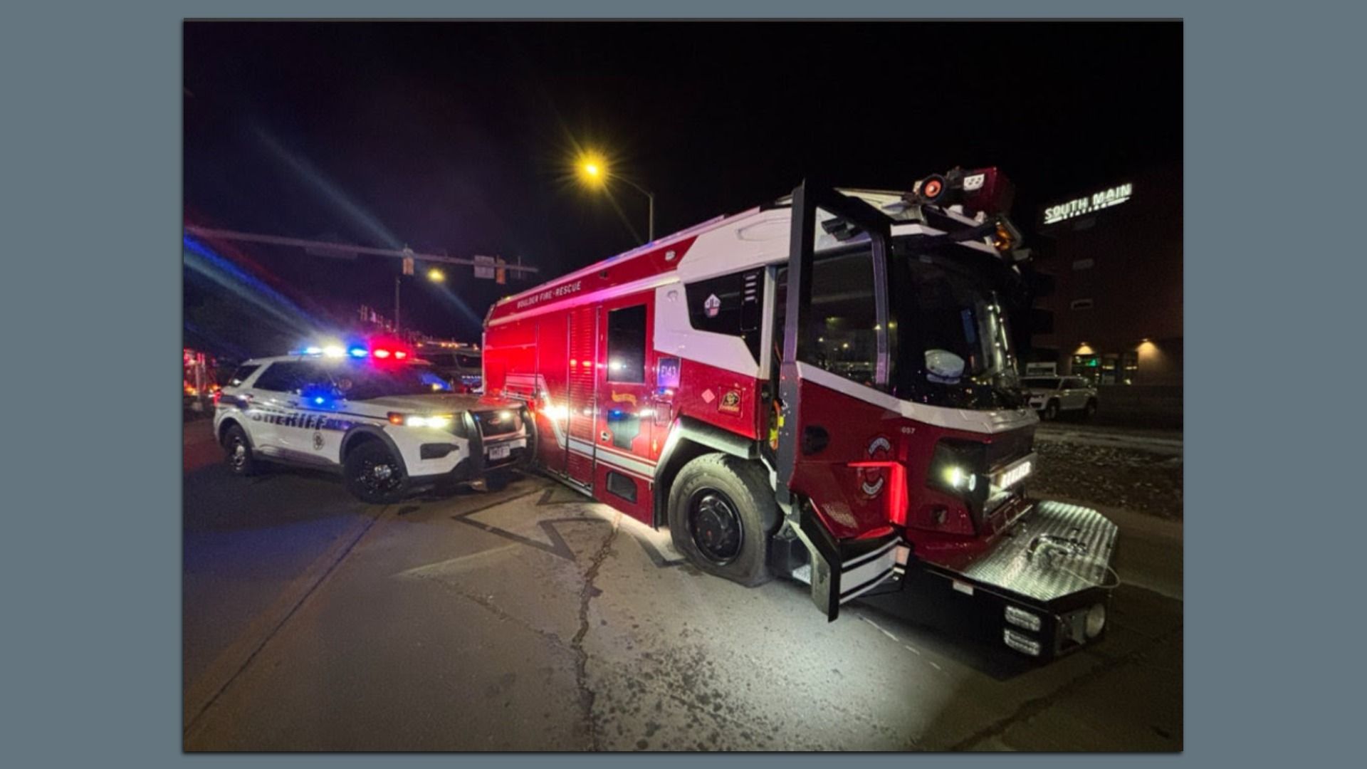 Night scene of a red and white fire/rescue truck tipped on a street, with a sheriff SUV and flashing blue and red lights nearby; a yellow streetlamp shines, and a South Main sign is in the background.