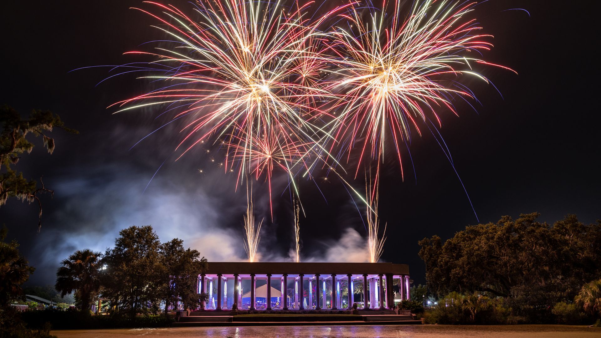 Photo shows a red, white and gold fireworks display exploding above the Peristyle, which is an open-air, columned building in City Park. It is night in the photo and trees are seen around the Peristyle.