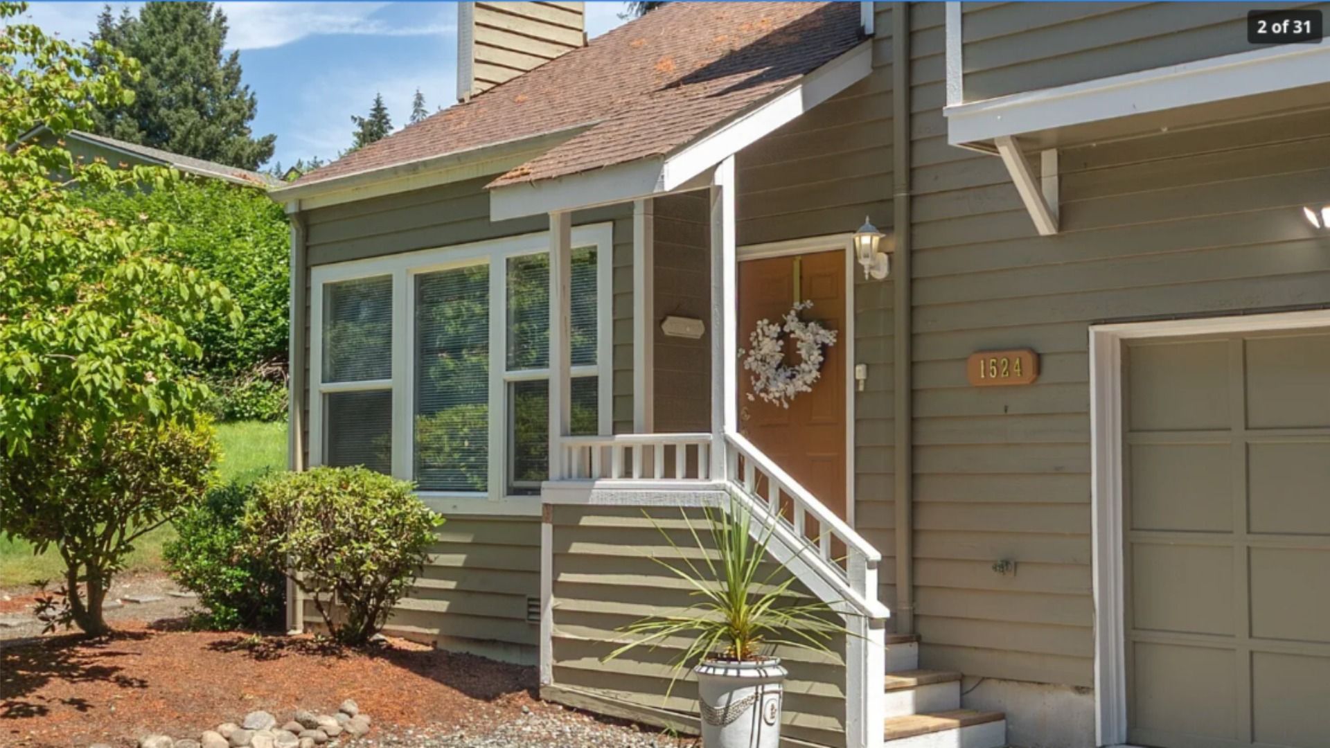 Front of a beige house with white trim. Brown door with a white wreath, small porch railing, and steps. Large window with blinds to the left, and a garage door to the right. Green shrubs and a tree.