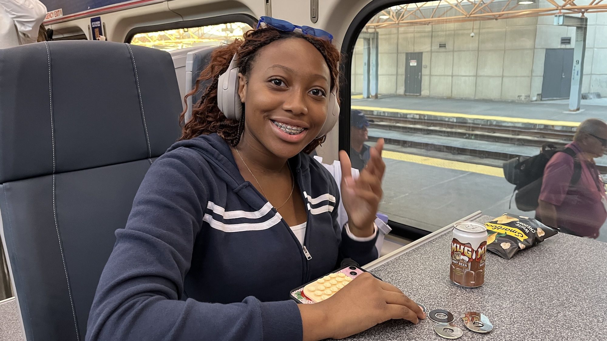 Laila Dunagan smiling with braces and red hair, wearing a navy hoodie and headphones, sits on a train with snacks and a can of Mug root beer on the table, with a station platform outside.