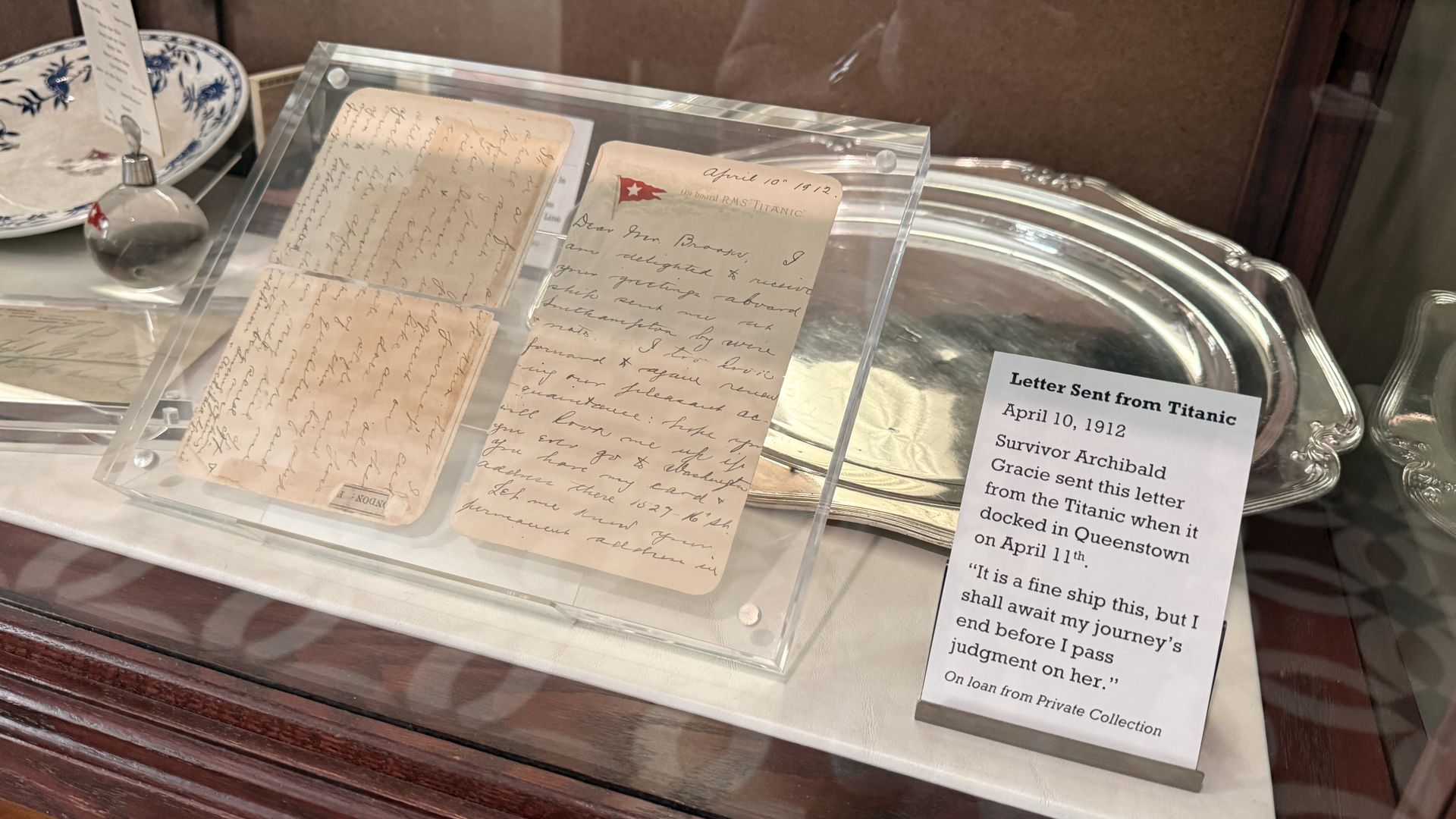 Glass display case showing two aged handwritten letters mounted in clear sleeves, a small red flag atop one page, with a silver tray in the back. A label reads "Letter Sent from Titanic" (1912).