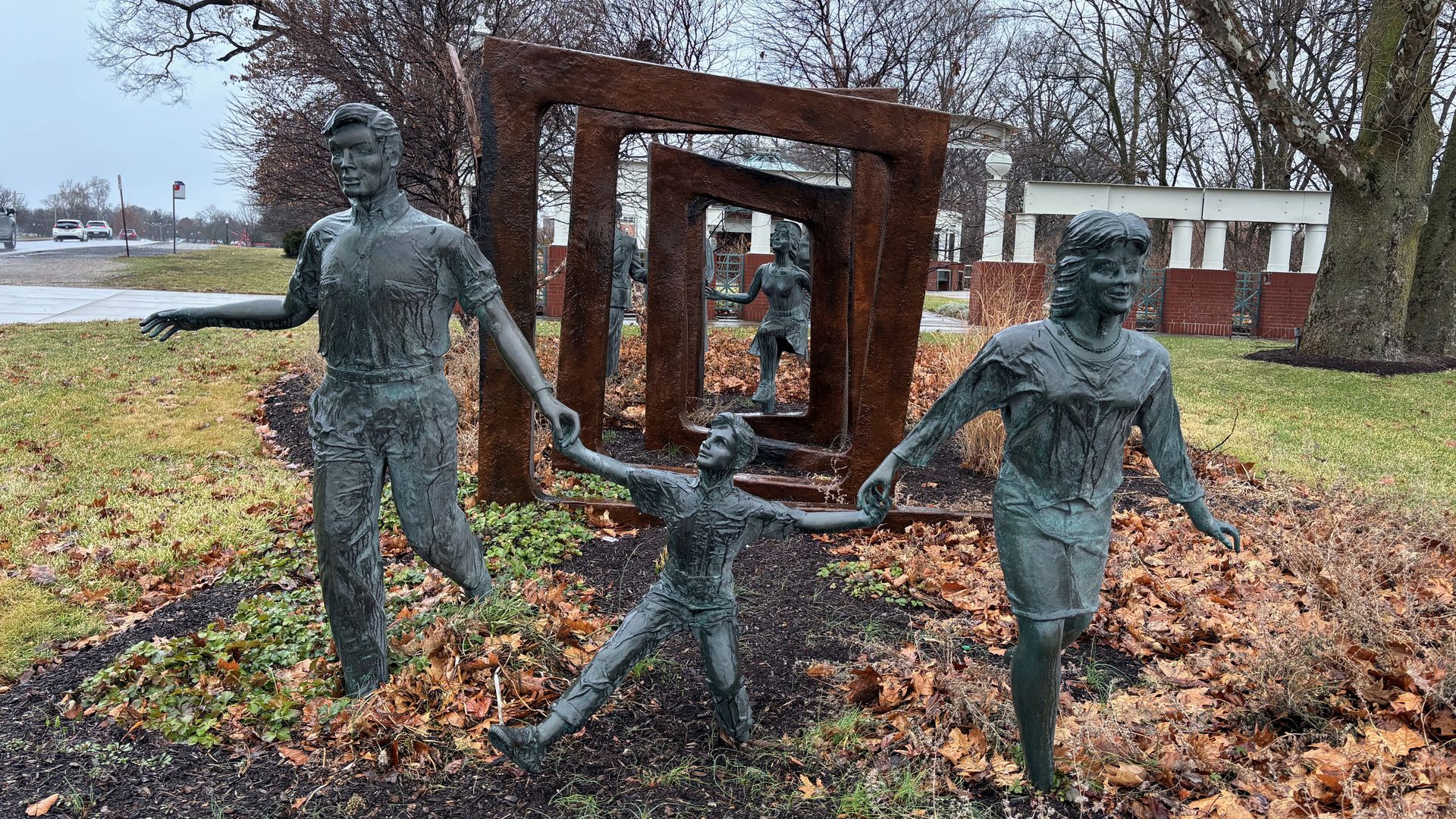 Bronze statues of a family with a man, child, and woman holding hands walking through a series of rust-colored rectangular frames in an outdoor park with bare trees on a gray day.