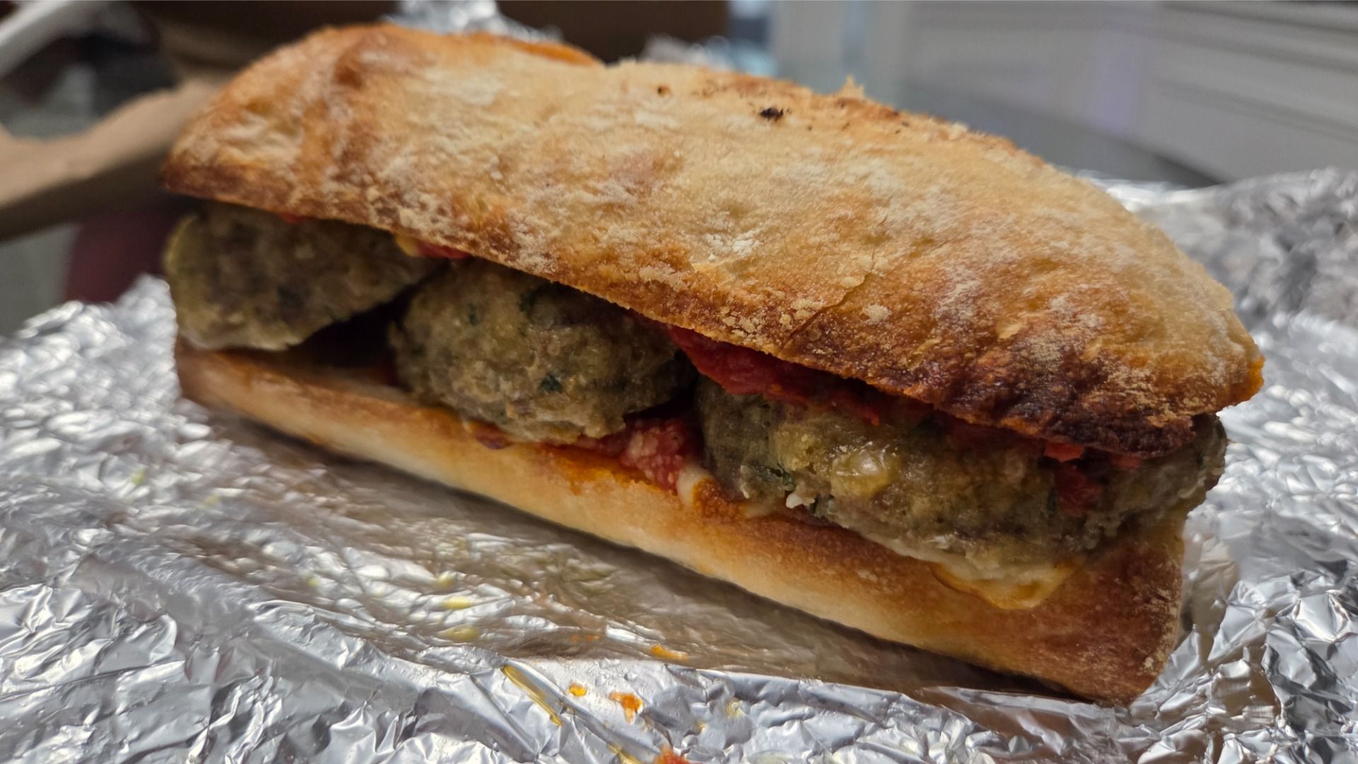 Meatball sandwich with toasted ciabatta bread and tomato sauce on foil, close-up showing three meatballs inside the sandwich.