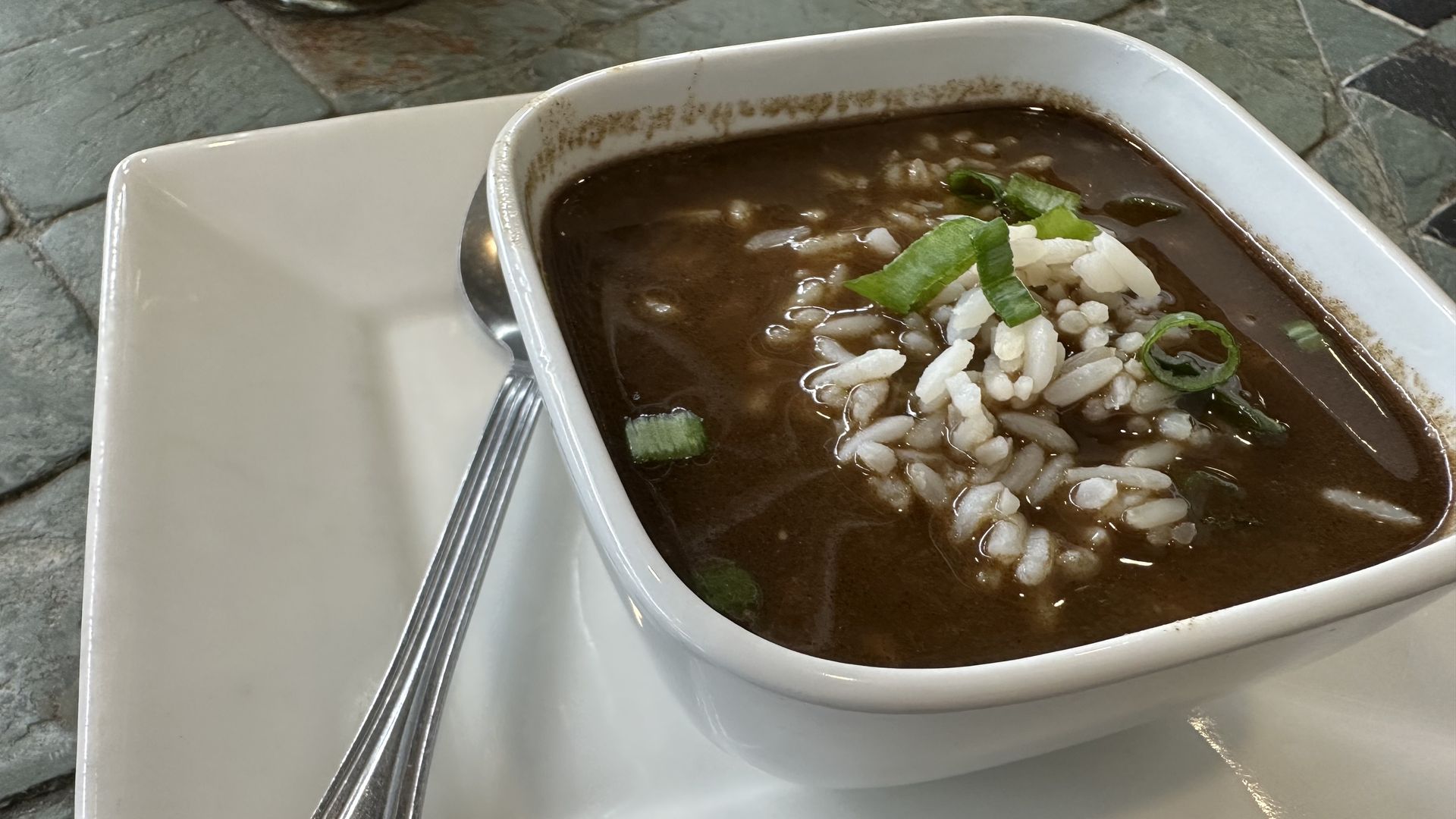 A bowl of gumbo sits on a plate with a spoon next to it. The gumbo has a dark roux and rice and green onions can be seen on top.