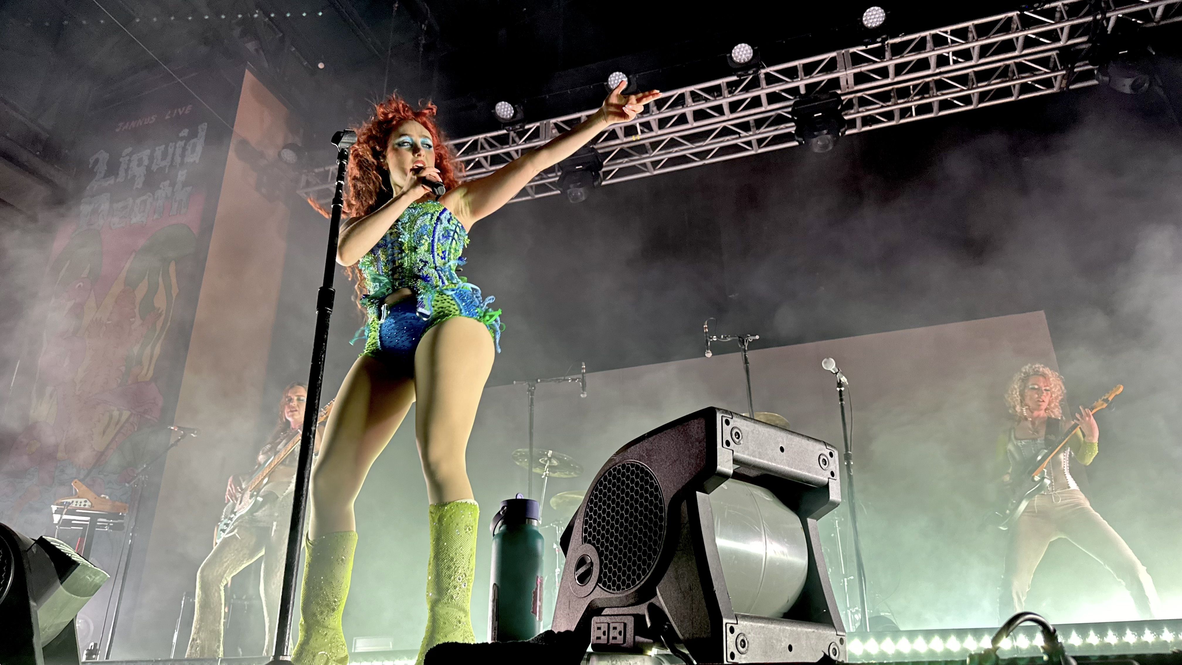 Chappell Roan onstage wearing a blue and green corset and green boots, pointing to the right