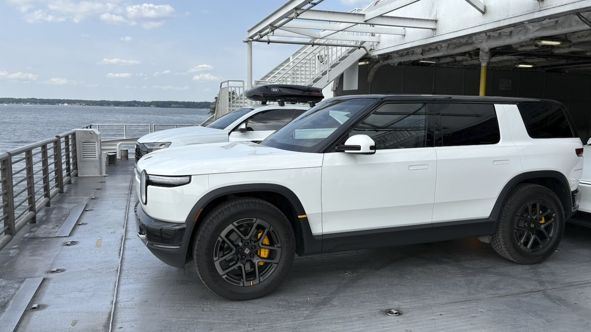 White Rivian R2 SUV with black wheels and yellow brake calipers parked on a ferry deck beside another white SUV; metal overhead beams, railing, and water with a blue sky in the background.