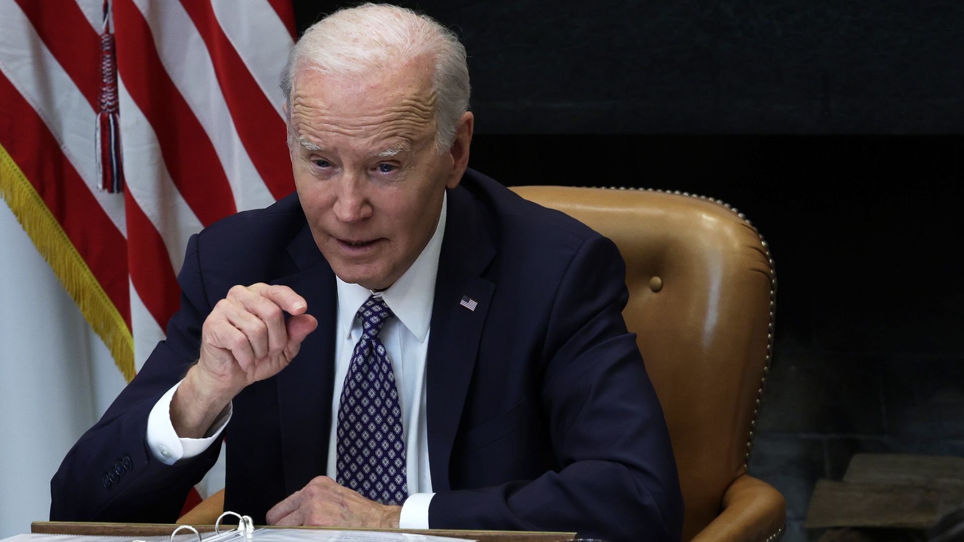 Joe Biden, wearing a blue suit and tie, and sitting in a leather chair, speaks to a group of people with an American flag in the background.