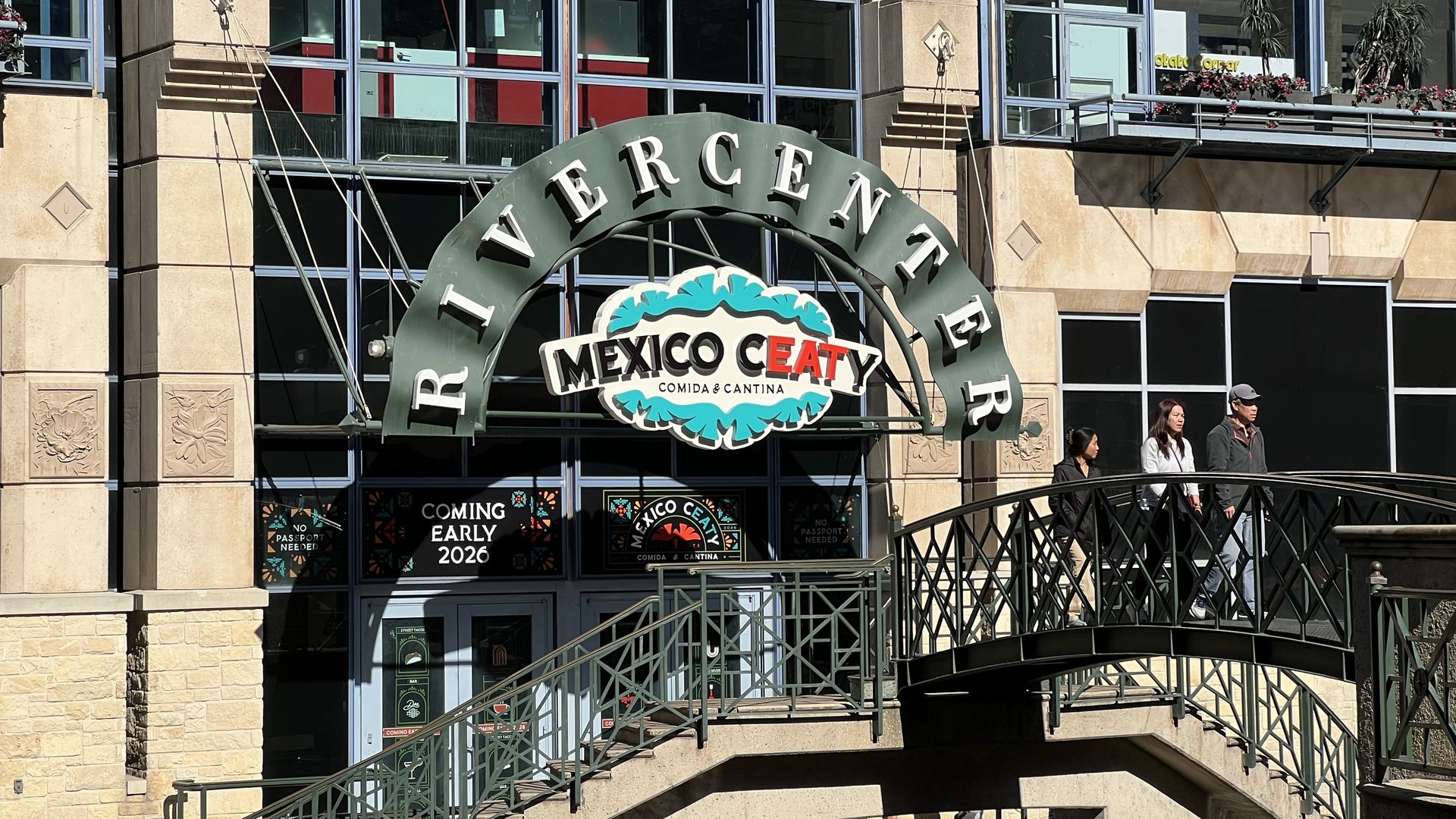 Green Rivercenter sign above entrance to soon-to-open Mexico Ceaty Comida & Cantina, with beige building facade and three people walking on a nearby bridge.