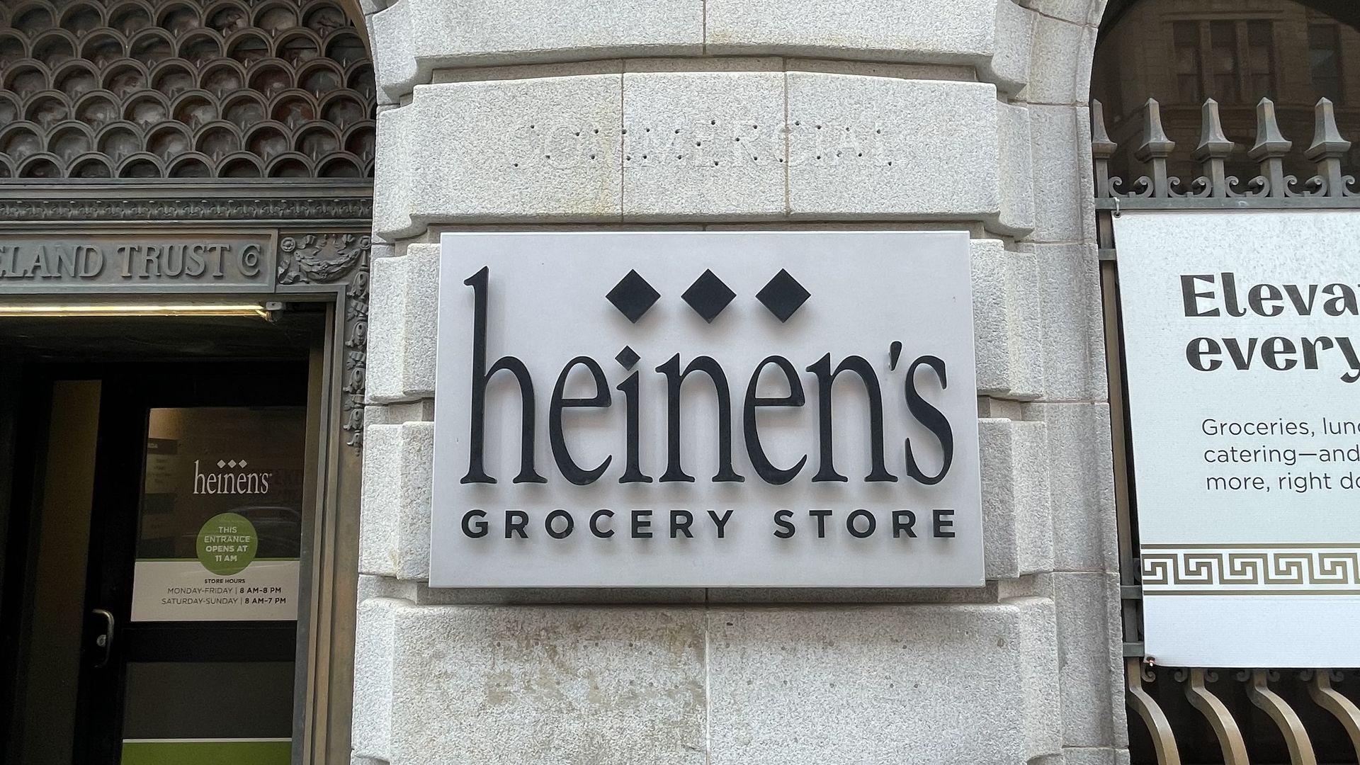 Gray stone storefront with a large white sign reading "heinens" in bold black letters, three black diamond shapes above, and "GROCERY STORE" below; ornate doorway on the left and railing on the right.