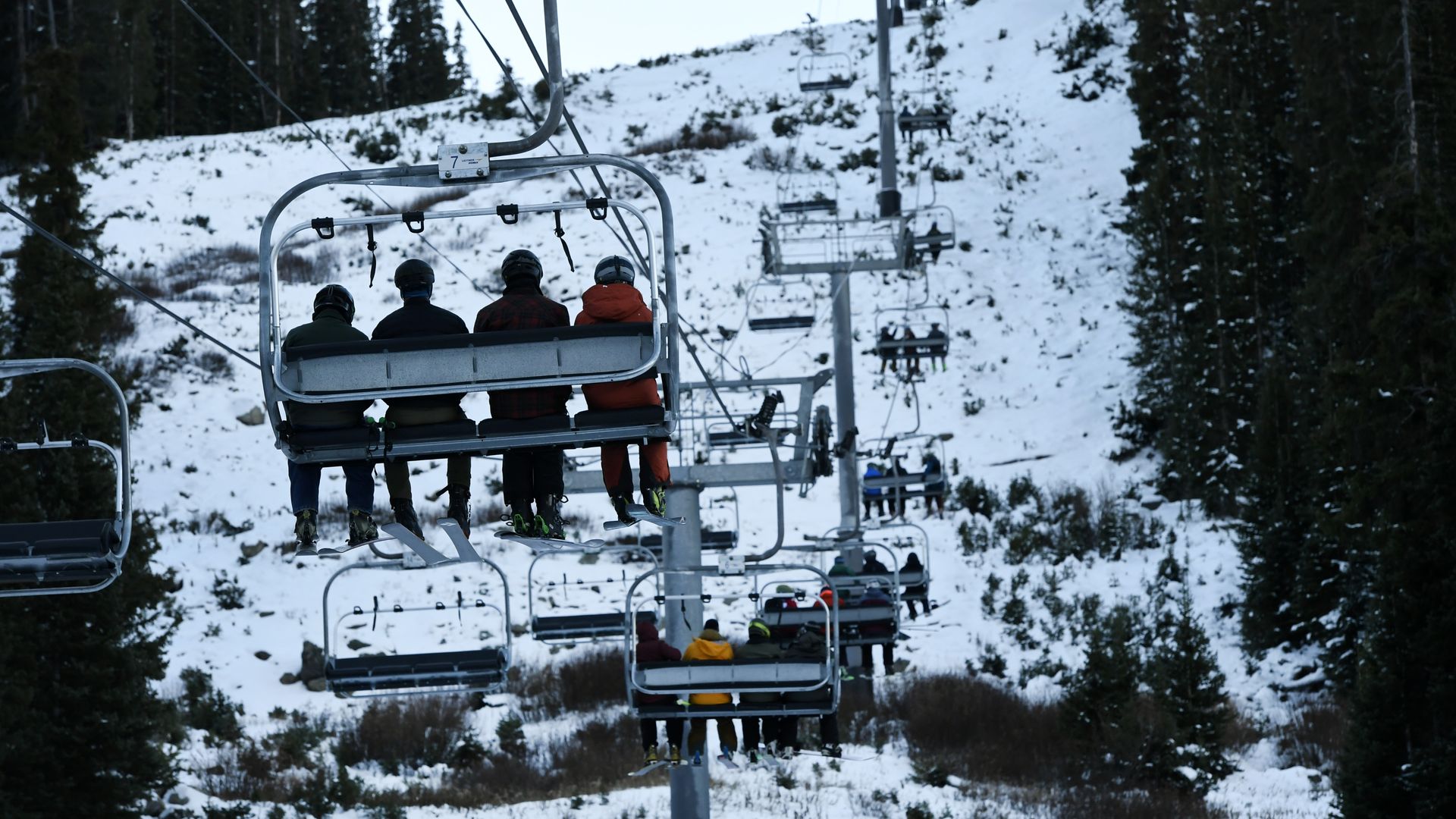 Four riders in helmets sit on a snow-covered ski-lift chair, one in an orange jacket; a row of chairs climbs the slope amid dark evergreen trees.