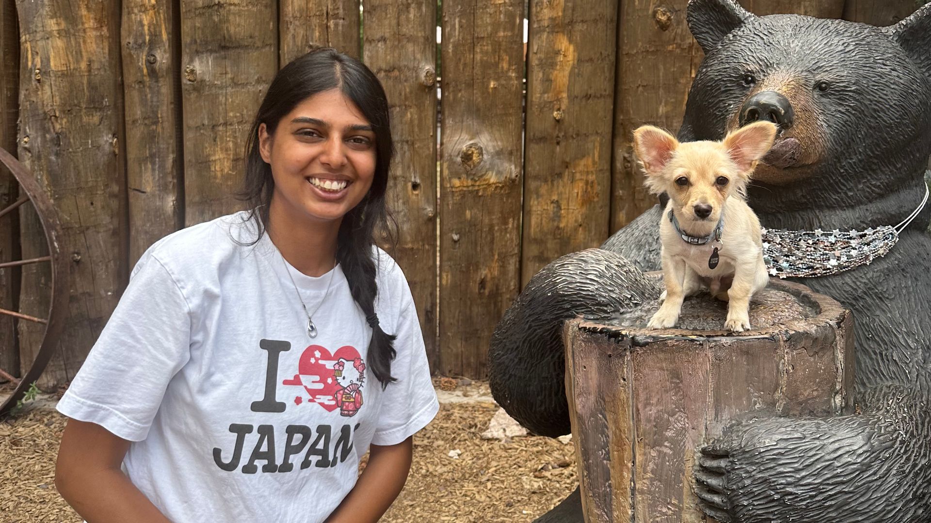 Smiling woman in a white T-shirt sits beside a large bear statue with a small tan dog on its arm; behind them is a rustic wooden sign reading "BEARIZONA" with a metal paw print.
