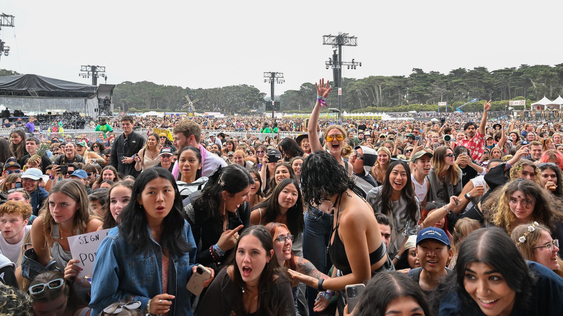 Large outdoor music festival crowd, stage with a black tent on the left, fans raise hands and phones, bubbles float above, forested trees in the background, sunny daytime.