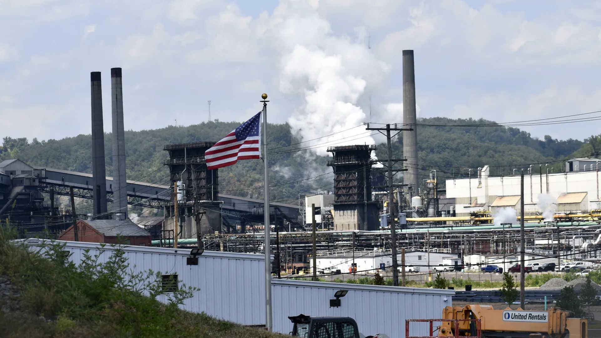 Industrial facility with tall smokestacks, complex piping, and machinery. An American flag flies on a pole in the foreground as white smoke pours from a chimney; blue sky with clouds behind hills.