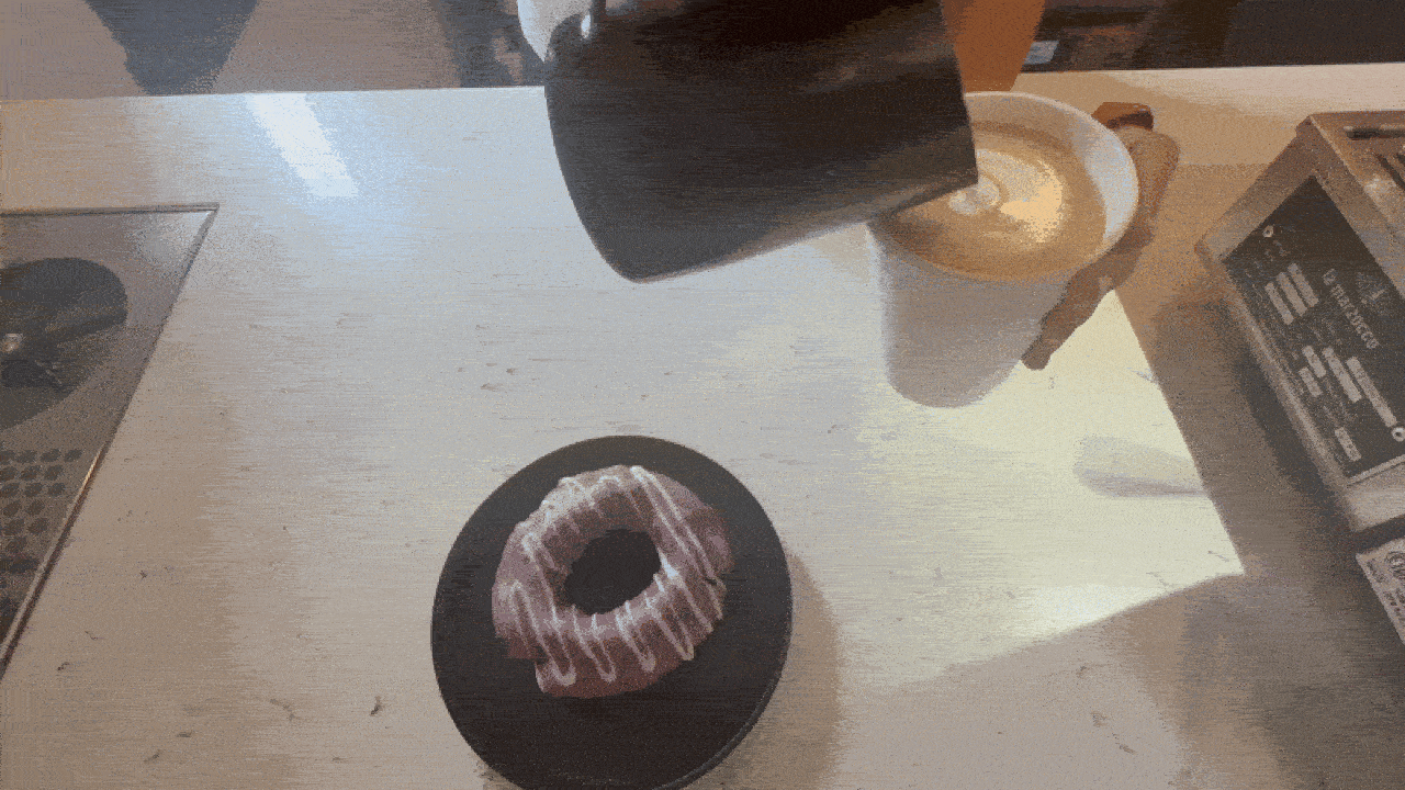 Person pouring milk into a cup to make latte art on a light wooden table with a pink-frosted donut with white icing drizzle on a black plate nearby.