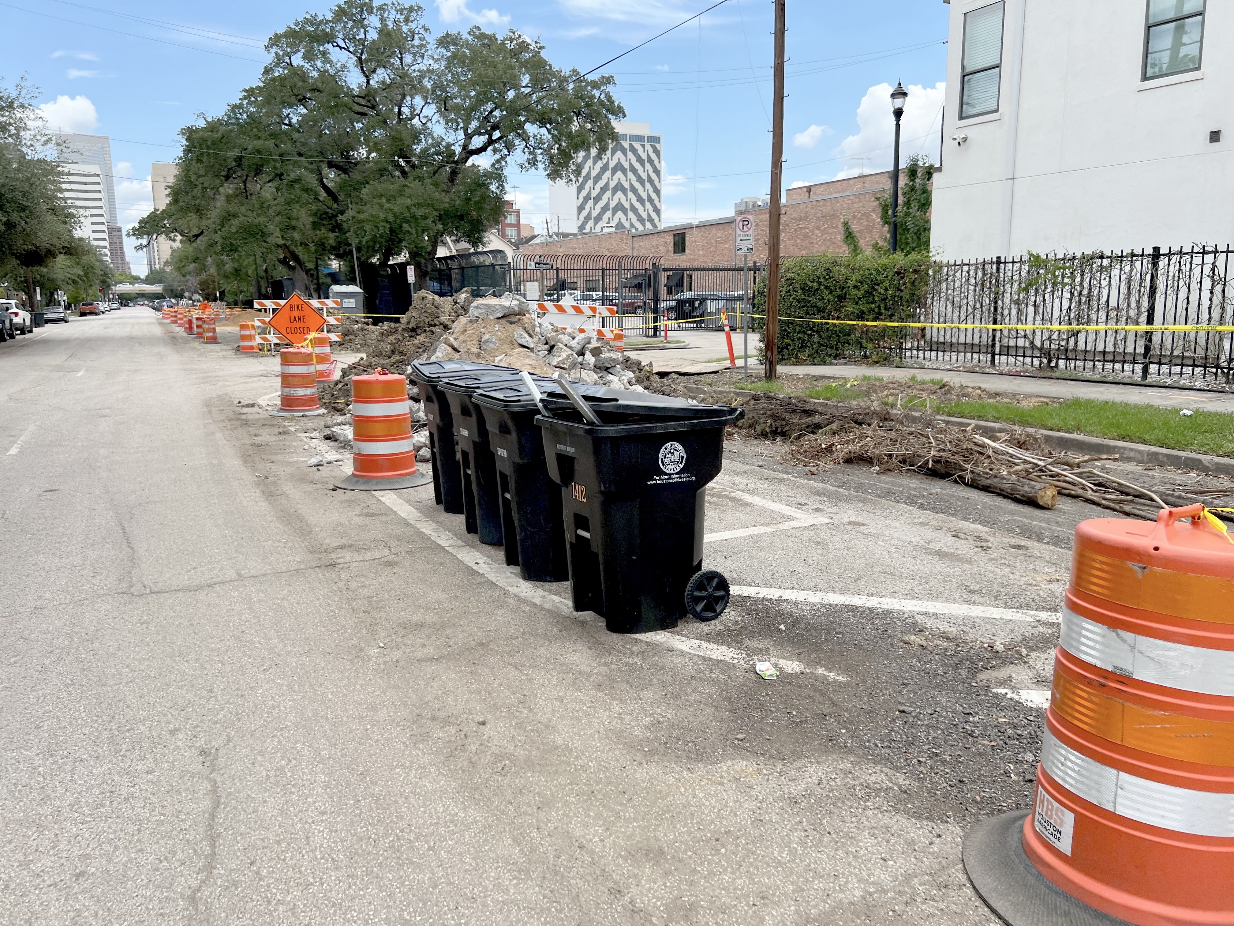 Street under construction with orange barrels, piles of rubble, fallen tree branches, black trash bins 