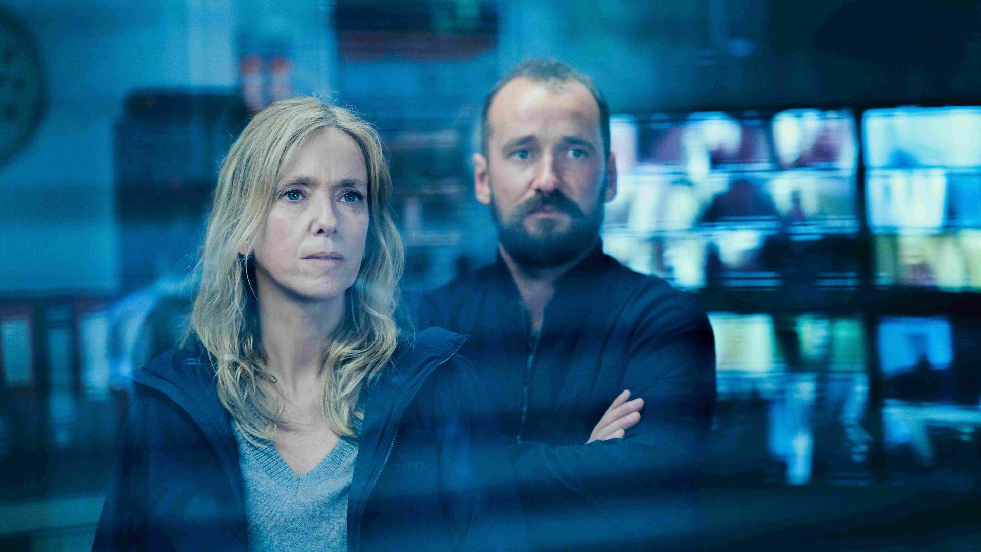 Two people in a blue-lit control room. Foreground: a serious blonde woman in a gray sweater; behind her, a bearded man in dark clothing with arms crossed as screens glow in the background.