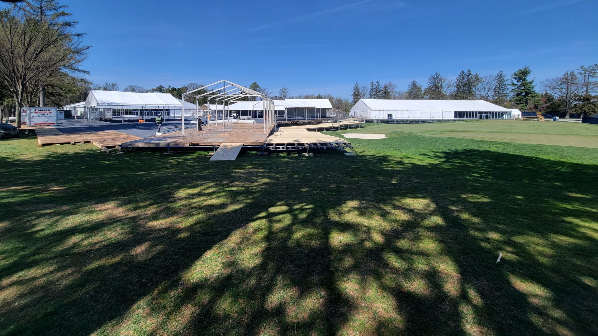 Sprawling tent structures, a wooden raised-stage platform, a metal-frame canopy at Aronimink Golf Club.