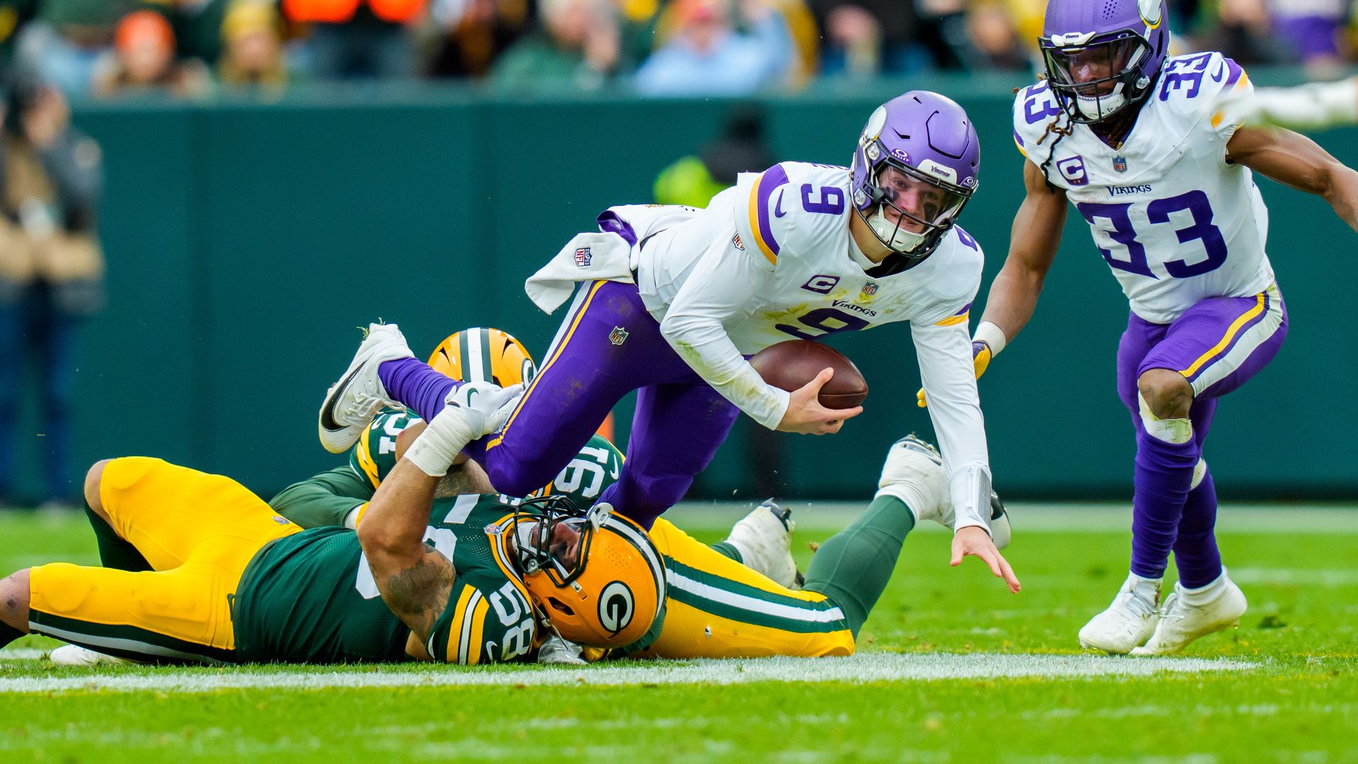 A football quarterback in purple and white Vikings uniform gets tackle by two Green Bay Packers players in green and yellow on a grassy field during game.
