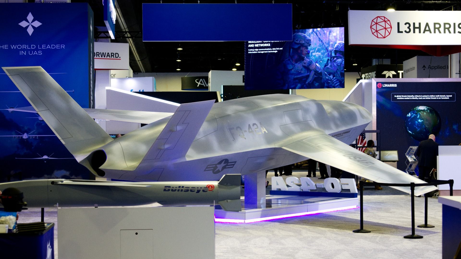 Large gray unmanned aircraft model on display at a defense expo. It's surrounded by defense contractor signs and advertisements.
