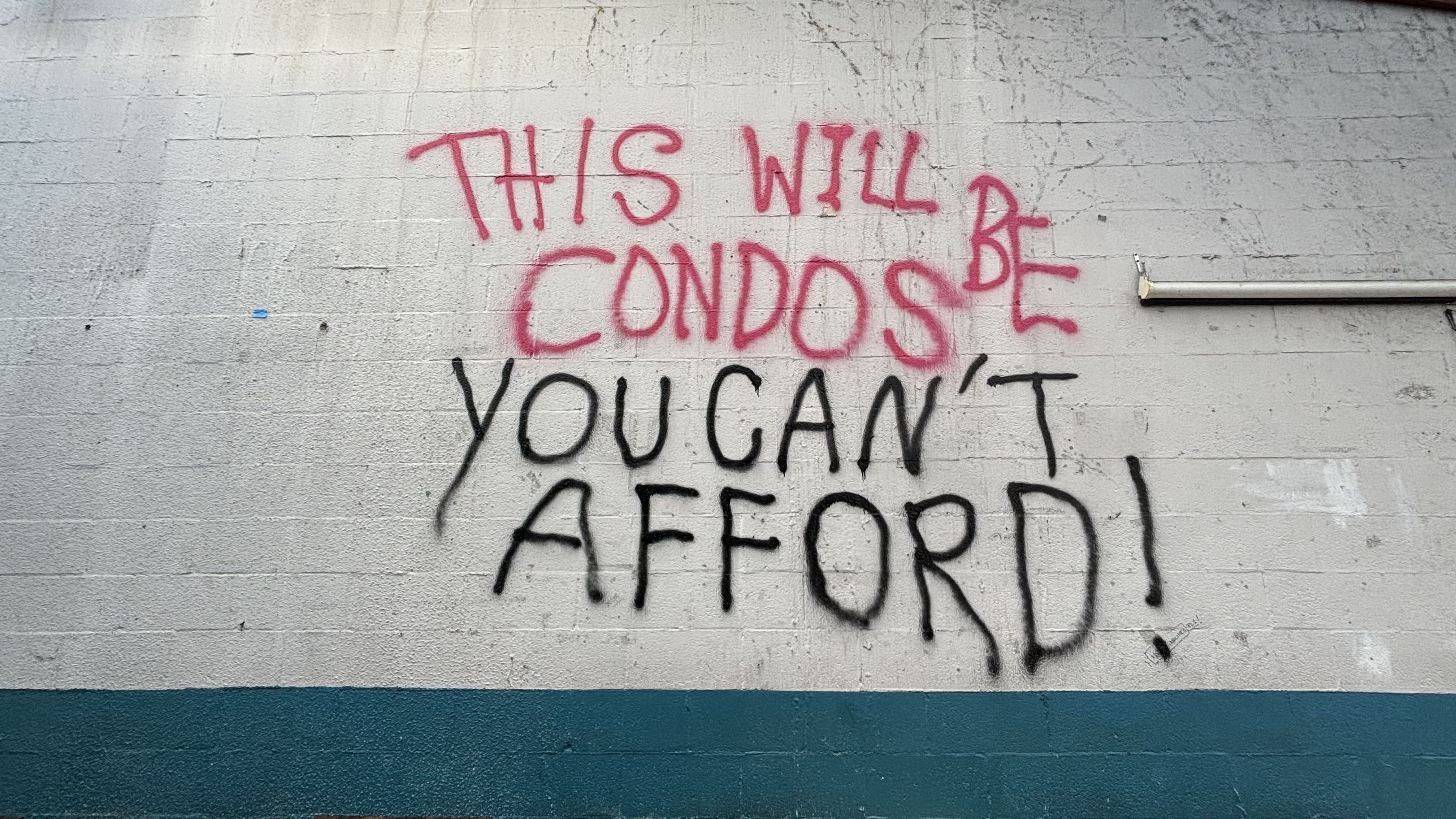 Graffiti on a white cinder block wall with teal trim reads in red and black spray paint: "THIS WILL BE CONDOS YOU CAN'T AFFORD!"