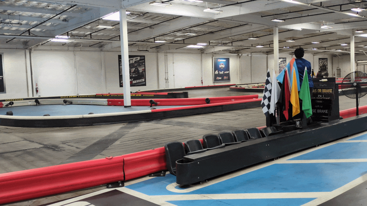 Indoor go-kart track with red and black safety barriers, winding gray asphalt track, a podium with checkered and multicolored flags, row of black seats, and a person standing near the flags.