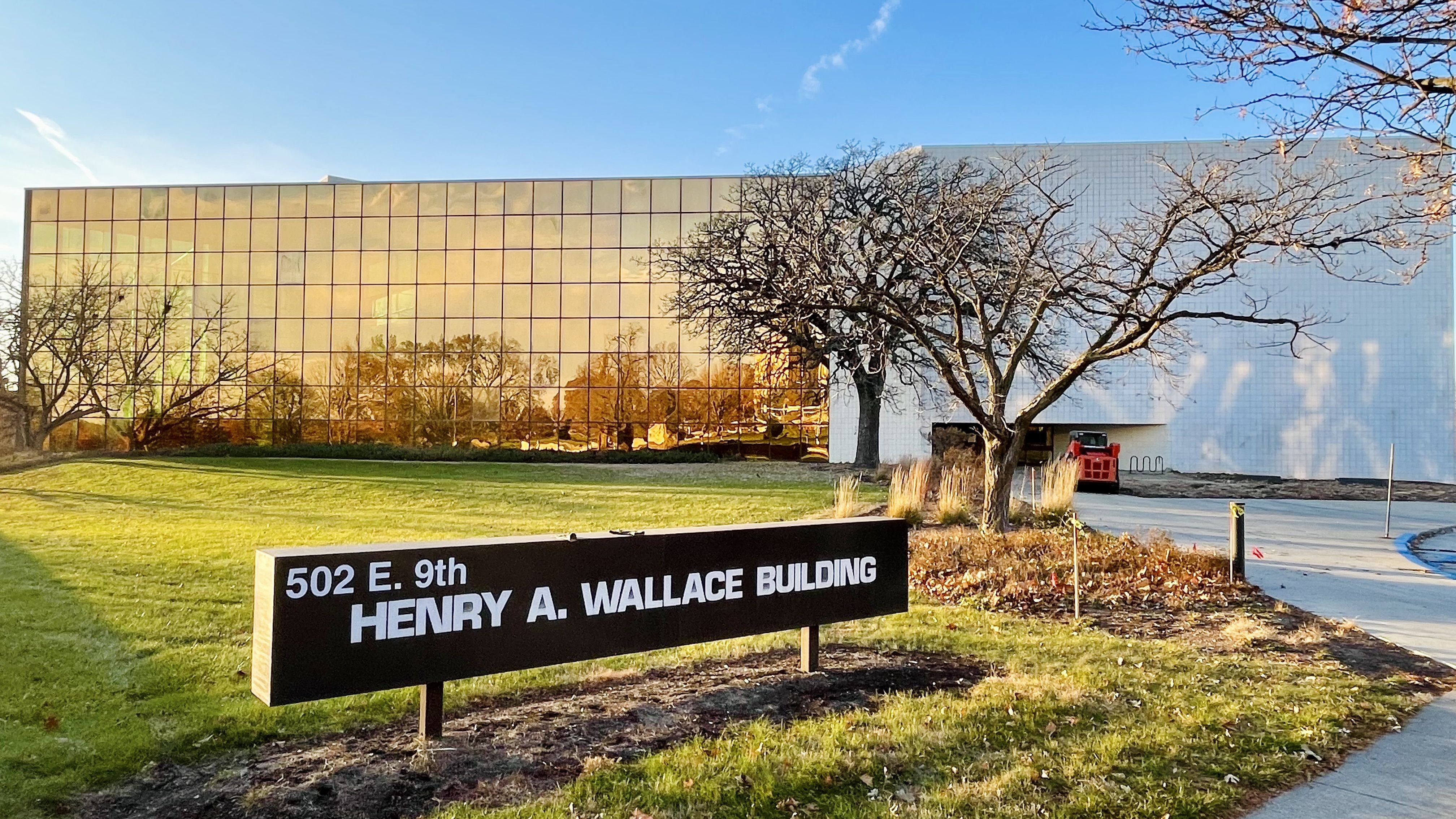Building with golden reflective windows and white wall, bare trees, green grass, and sign reading 502 E. 9th HENRY A. WALLACE BUILDING under clear blue sky.