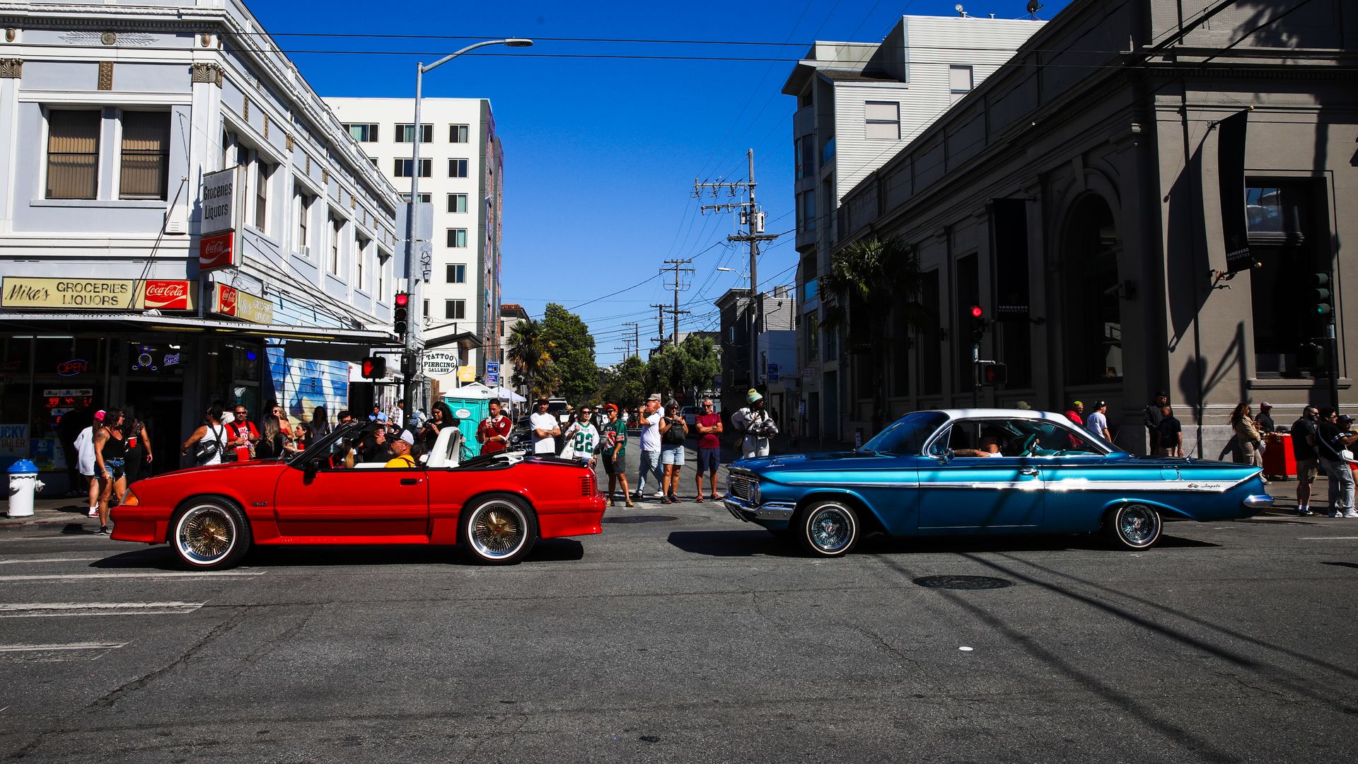 Red convertible and blue vintage car stopped at intersection with crowd watching outside a grocery and liquor store under clear blue sky.