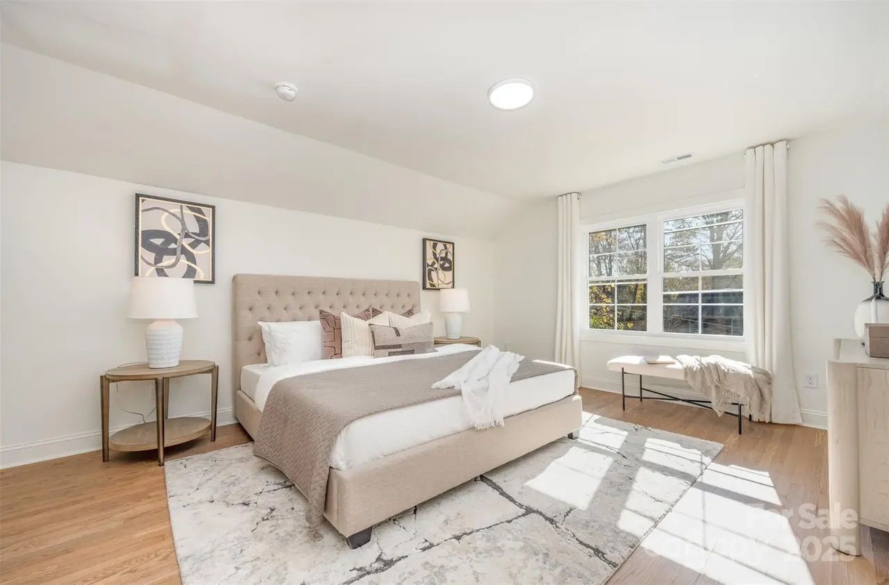 Bright bedroom with beige upholstered bed, white and brown bedding, two side tables with lamps, abstract wall art, large window with white curtains, wooden bench, and light hardwood floor.