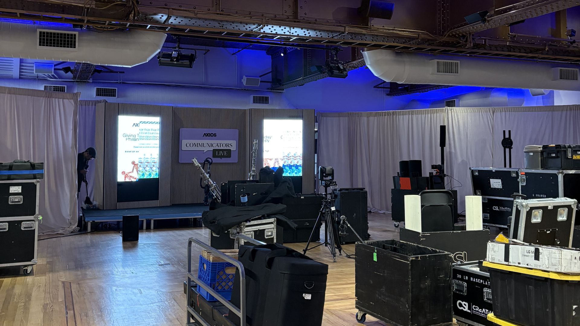 Event setup in a room with wooden floor and exposed brick ceiling lit with blue lights. Various equipment cases and carts are scattered around near a stage with screens displaying "AXIOS COMMUNICATORS LIVE."