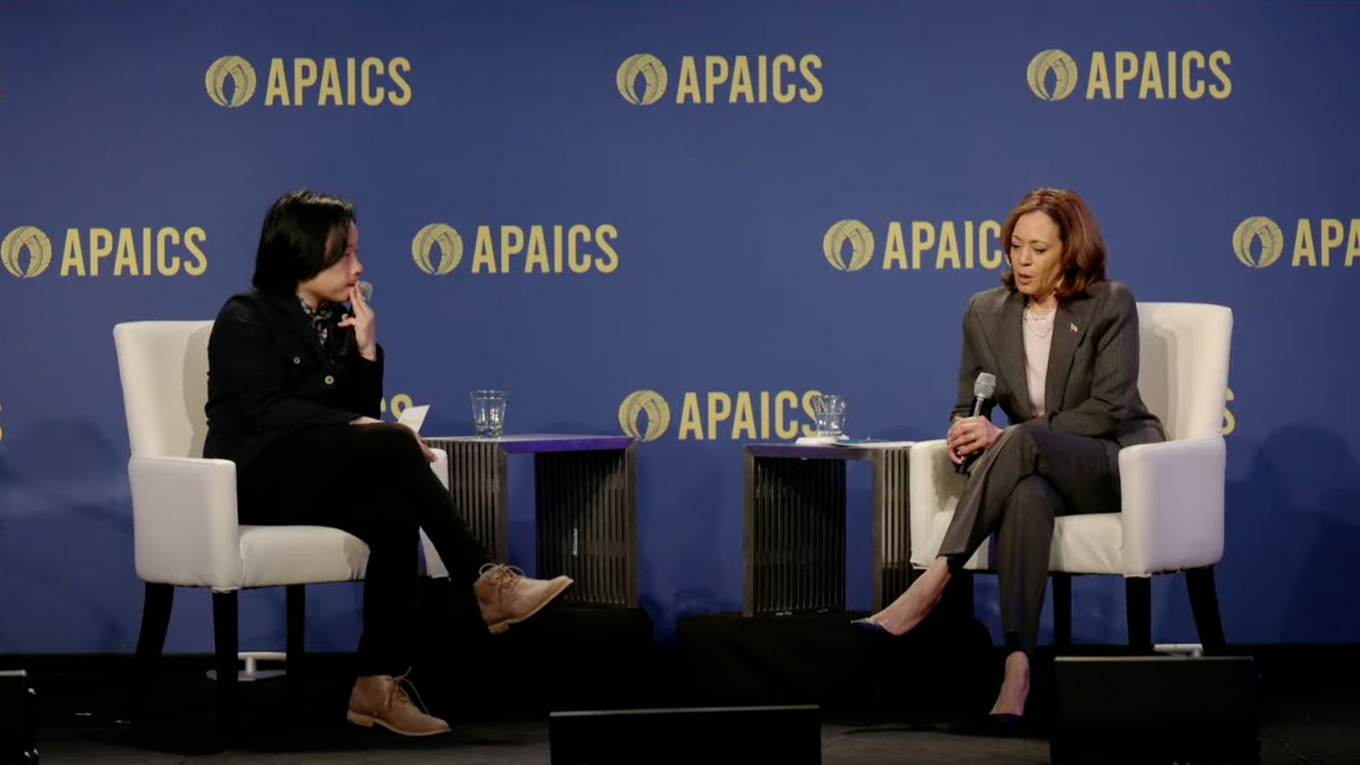 Kamala Harris, right, sits during an interview panel. Behind the seats is a backdrop that says APAICS repeatedly.