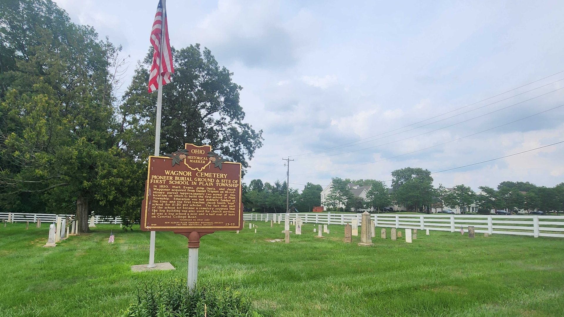 Ohio Historical Marker at Wagner Cemetery near a flagpole with a U.S. flag, surrounded by green grass, several old gravestones, a white fence, and trees under a cloudy sky.