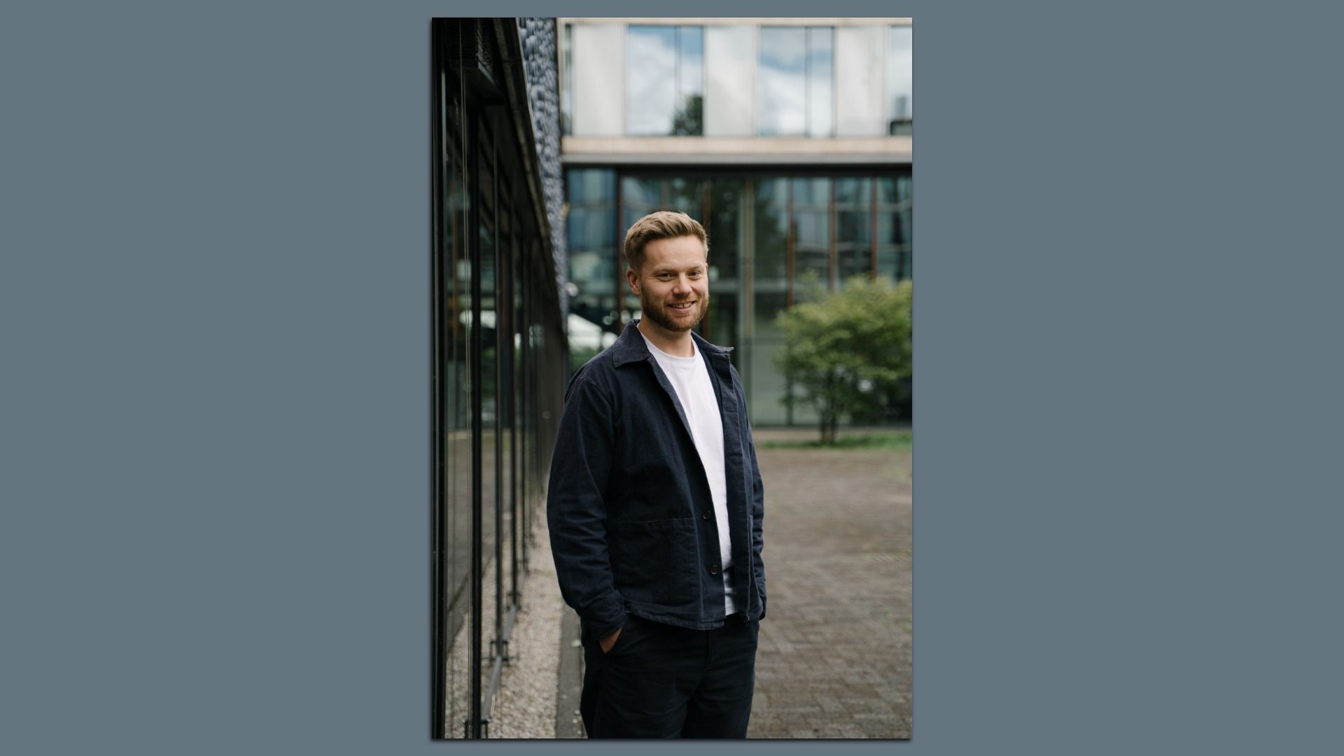 Smiling man with blond hair and light beard stands outside a modern glass building, wearing a white shirt and dark jacket, with greenery visible in the background.